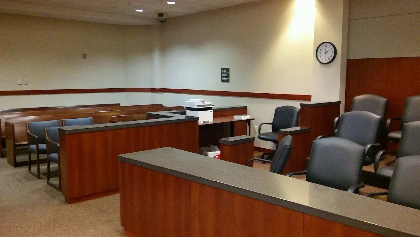 Empty courtroom with wooden benches, gray chairs, and a wall clock on beige walls