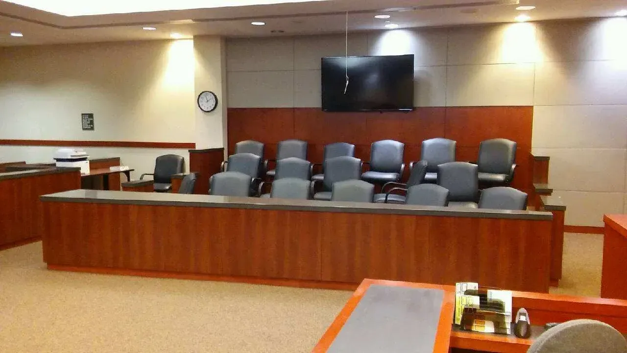Empty courtroom with wood-paneled judge’s bench, several gray chairs, and a wall-mounted screen