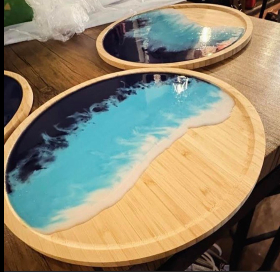 Oval wooden trays with blue resin ocean-wave designs, one in the foreground and one behind it
