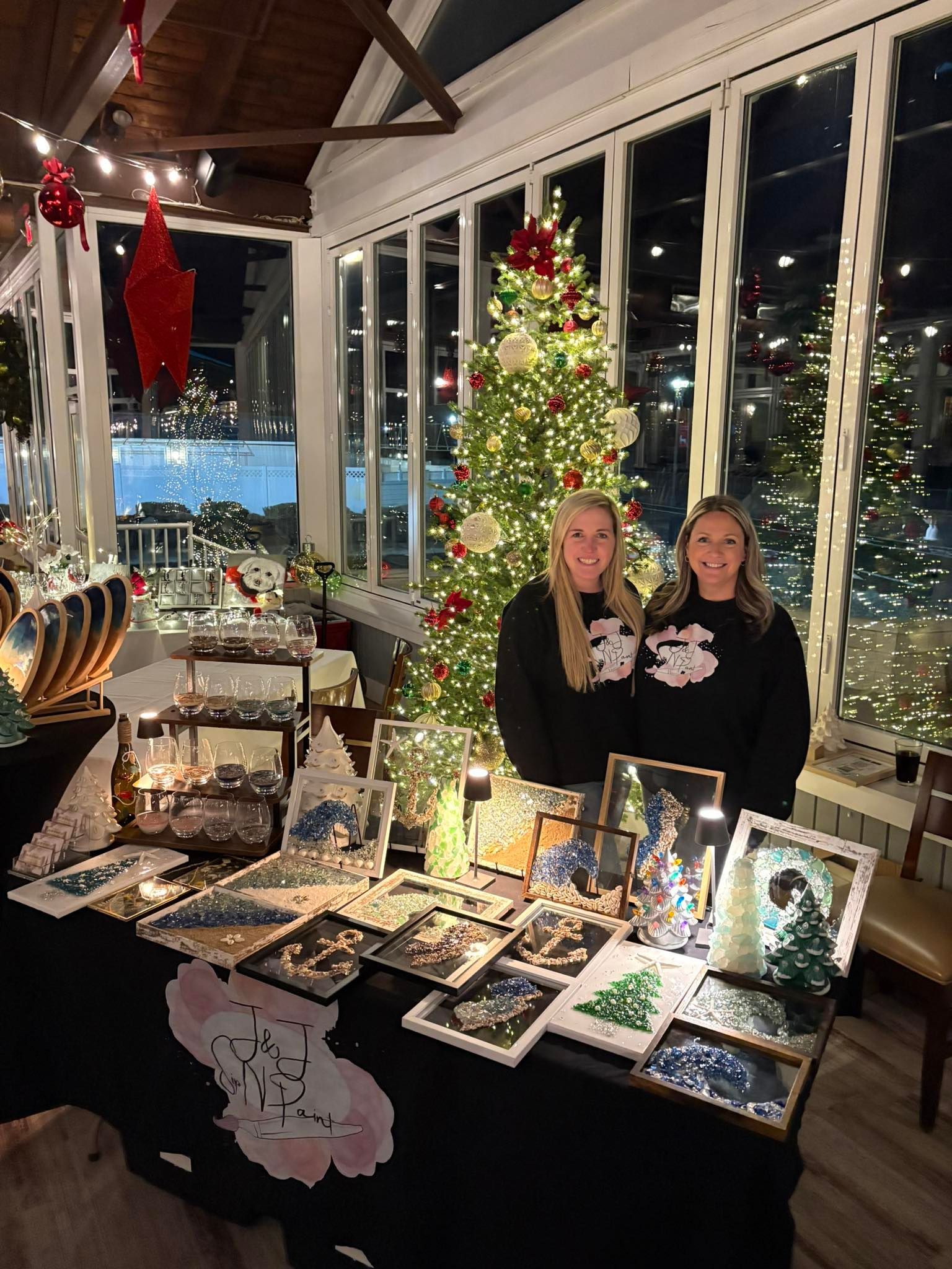 Two people behind a holiday craft display with lit Christmas tree and handmade cards in a restaurant window scene
