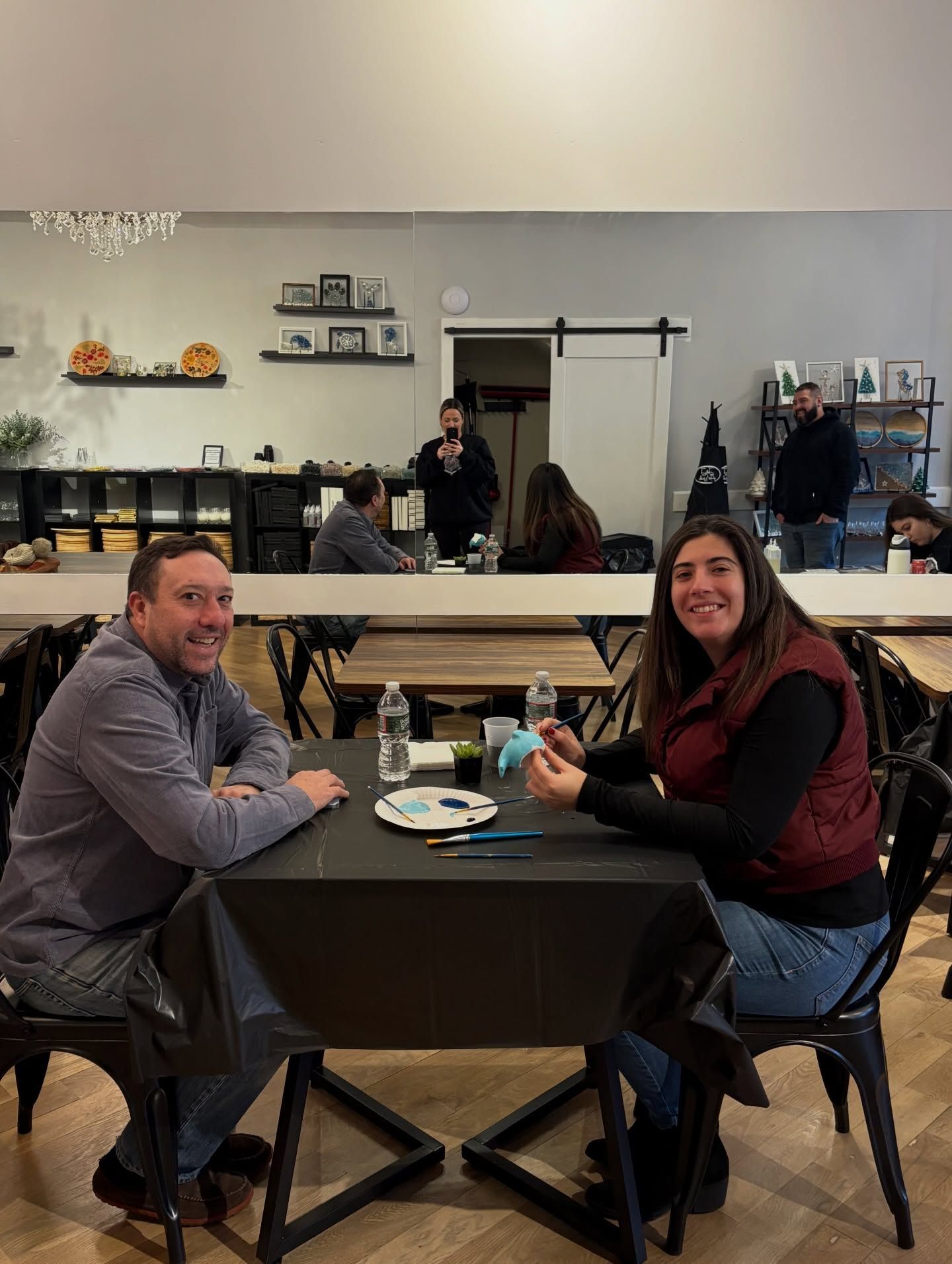 Two people smiling at a small café table in a rustic coffee shop, holding drinks and facing the camera.