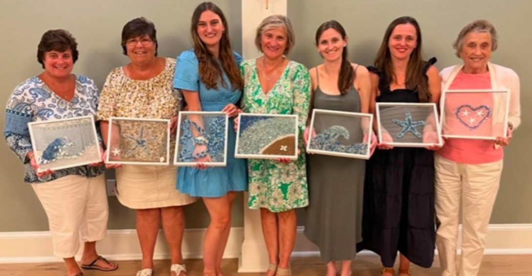 Seven people stand indoors holding framed artwork in a row, posing and smiling.