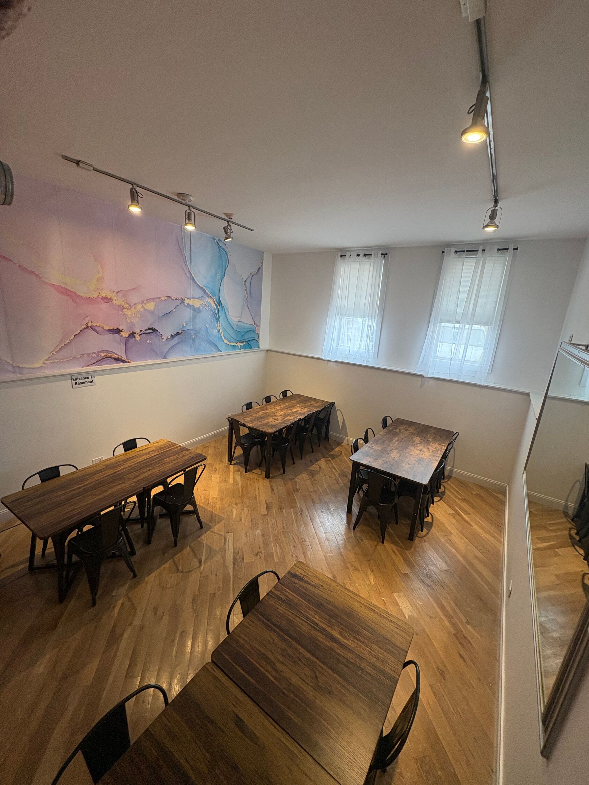 Empty modern dining room with wooden tables, black chairs, and large abstract wall art under track lighting.
