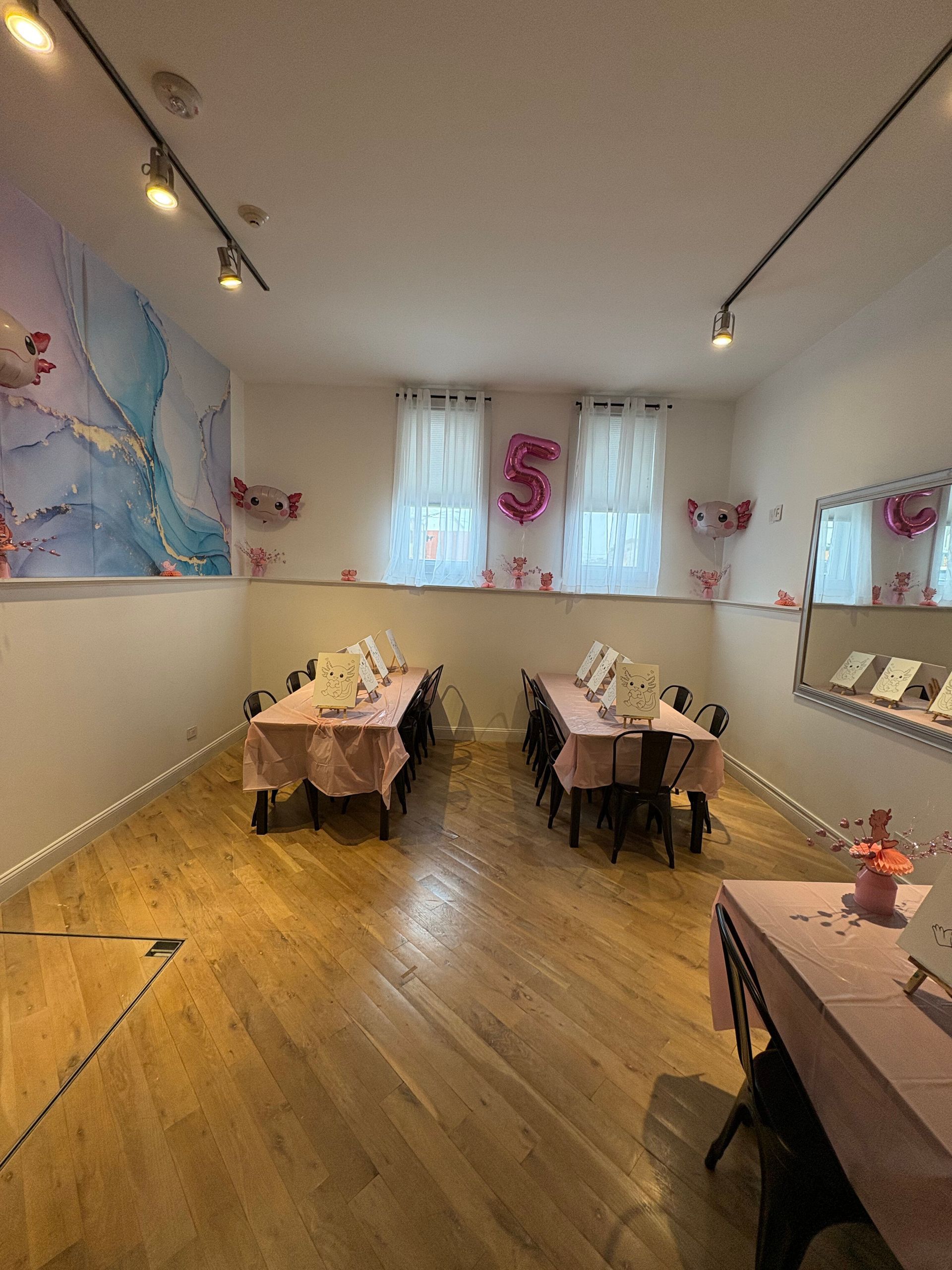 Small dining room with two set tables, wooden floor, track lighting, and soft wall art by windows.
