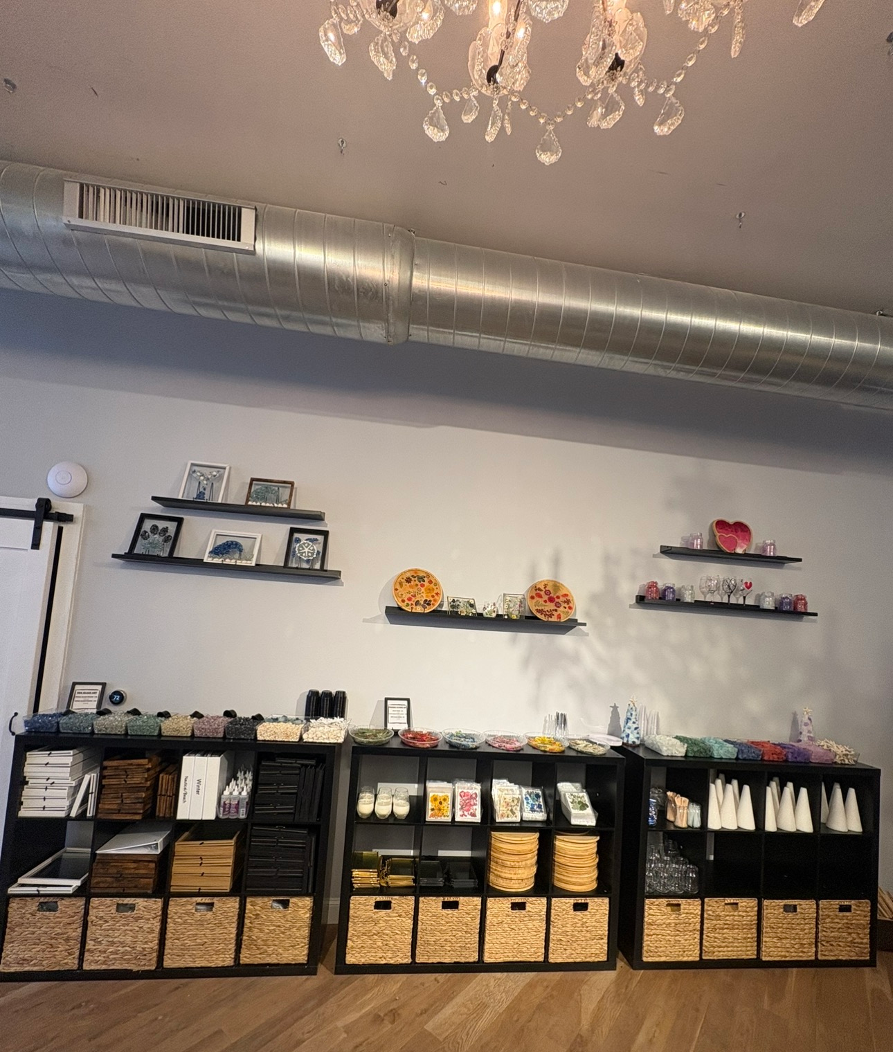 Retail display wall with shelves of small products, baskets, and wall decor beneath a ceiling vent and chandelier.