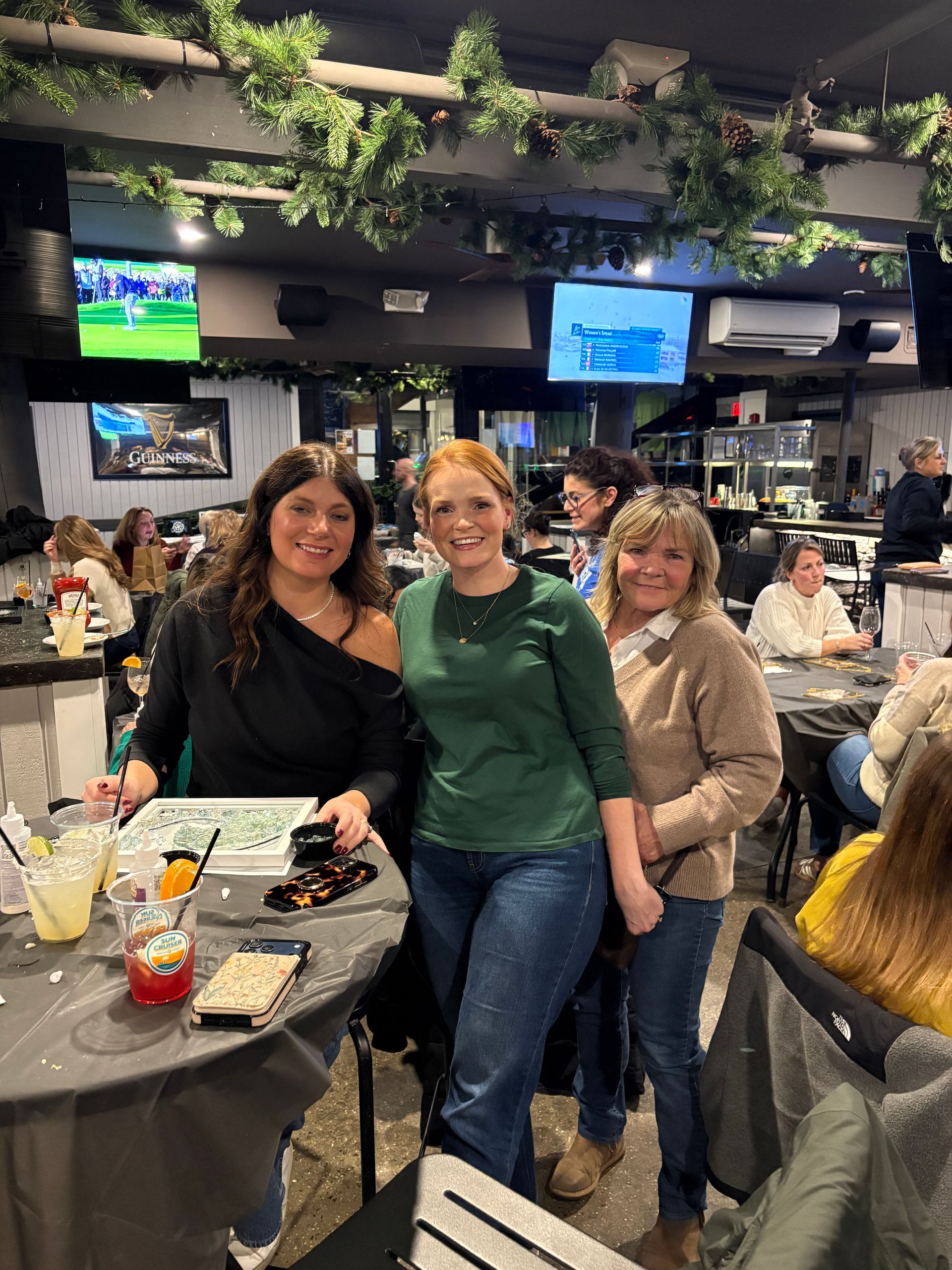 Three people posing at a lively bar, with drinks on a table and TVs overhead.