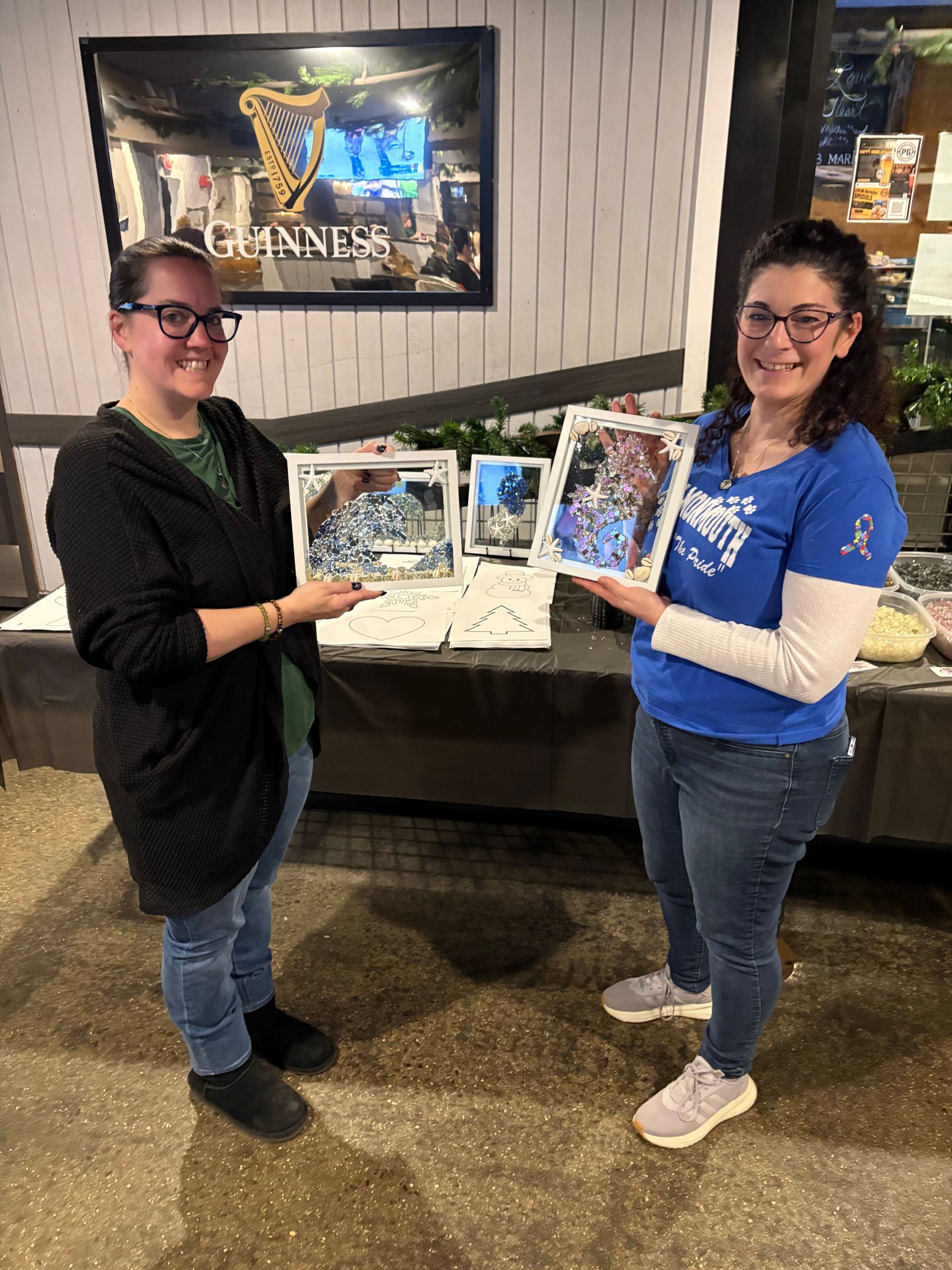 Two people holding up framed artwork at an indoor event booth with posters and a table behind them.