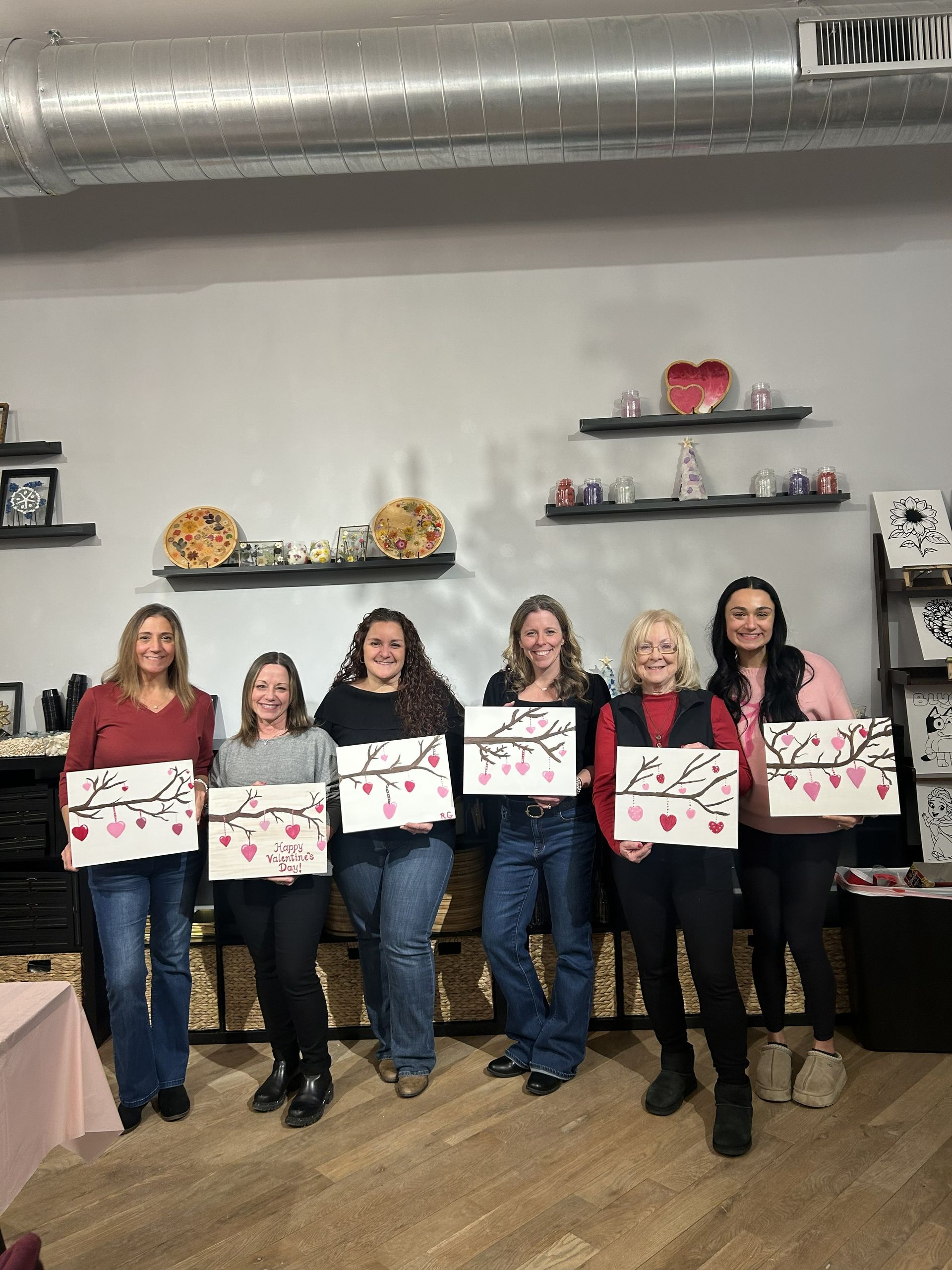 Six women stand indoors holding decorated square boards, posing in a room with wall shelves and a duct overhead.
