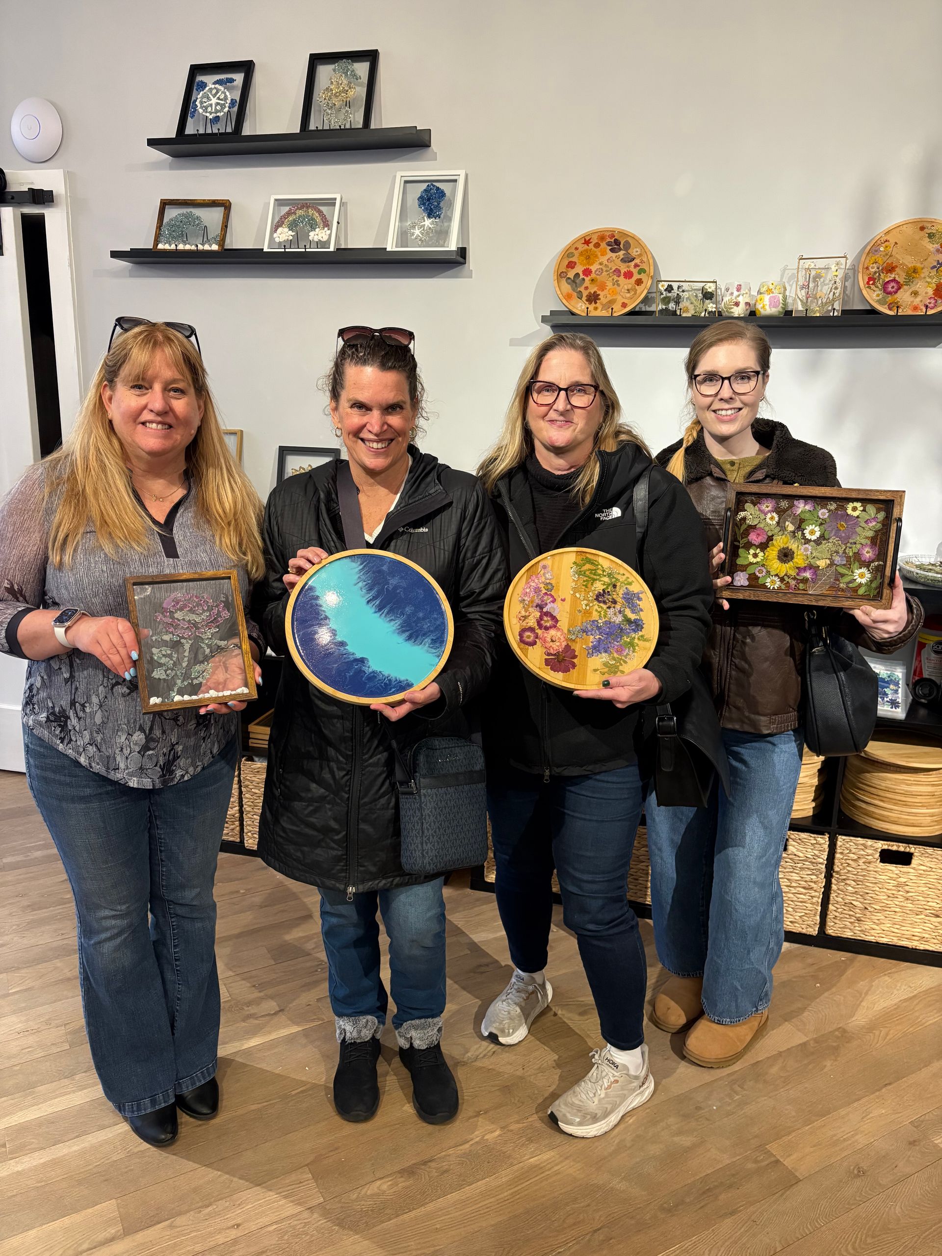 Four women holding colorful handmade art pieces in a craft studio, smiling in front of wall shelves.