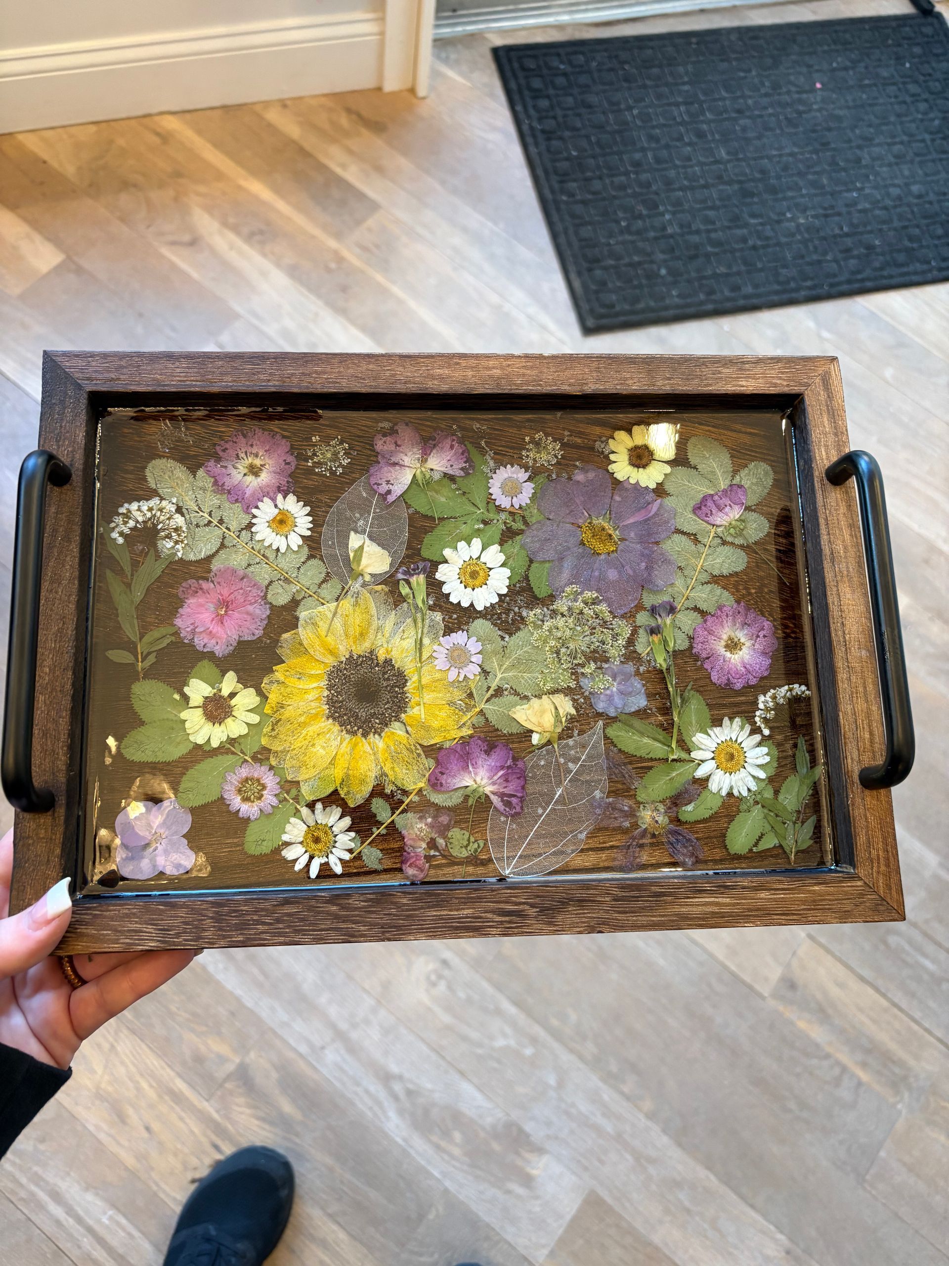 Wooden tray of pressed wildflowers with black handles on a light wood floor