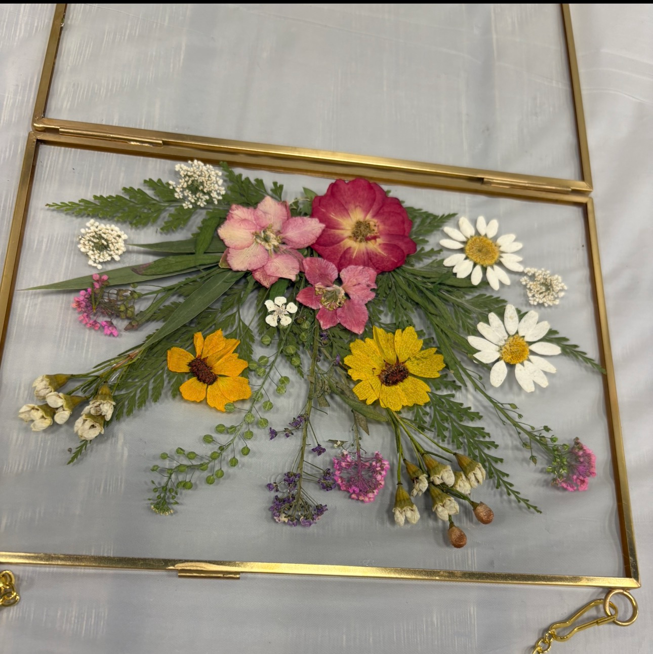 Pressed wildflowers arranged in a gold-framed display with pink, yellow, and white blooms on a light background