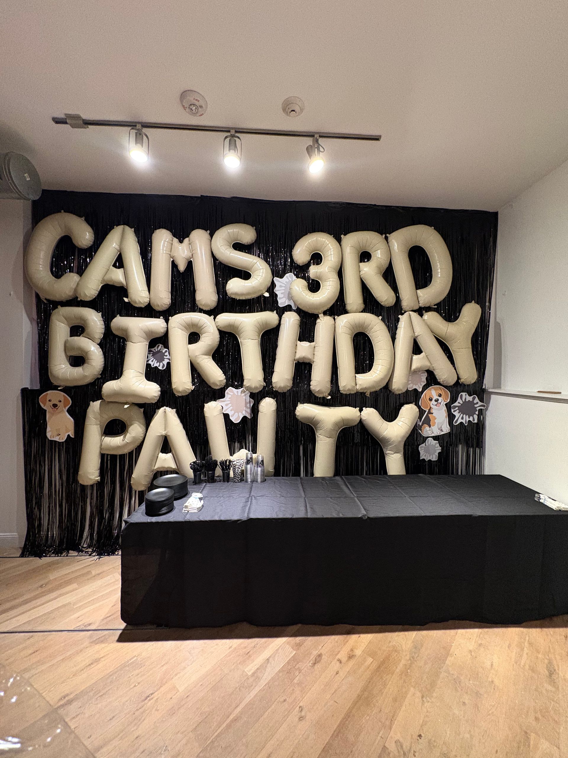 Birthday party setup with black table and large gold balloon letters reading “CAMS 3RD BIRTHDAY PARTY”