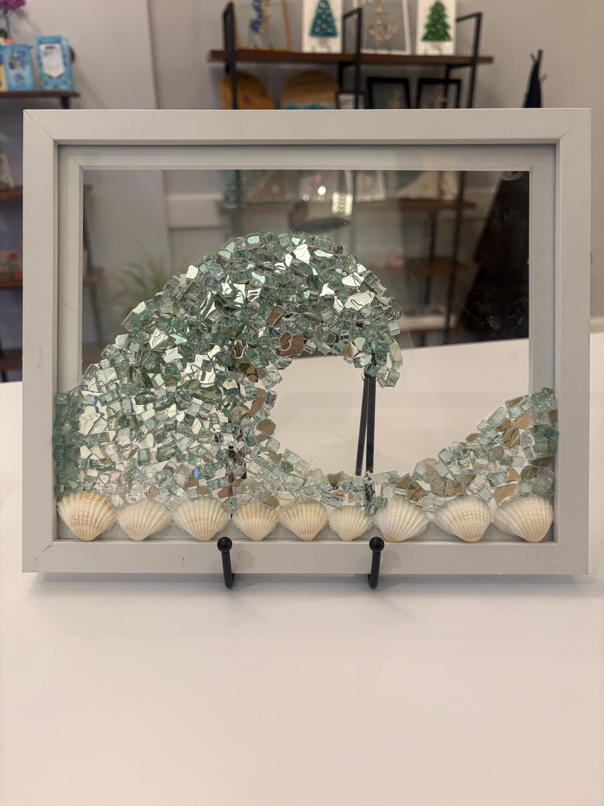 Framed glass artwork with a teal wave and white pebbles on a display stand