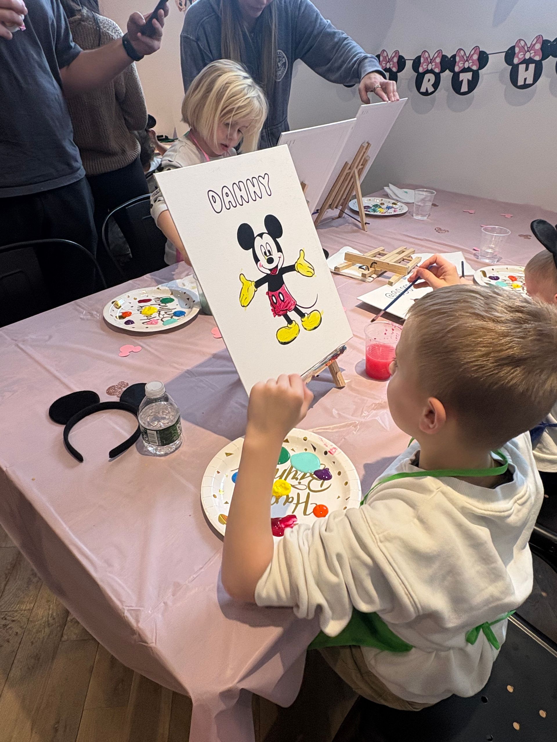 Child painting a Mickey Mouse craft at a table with paint palettes and paper decorations