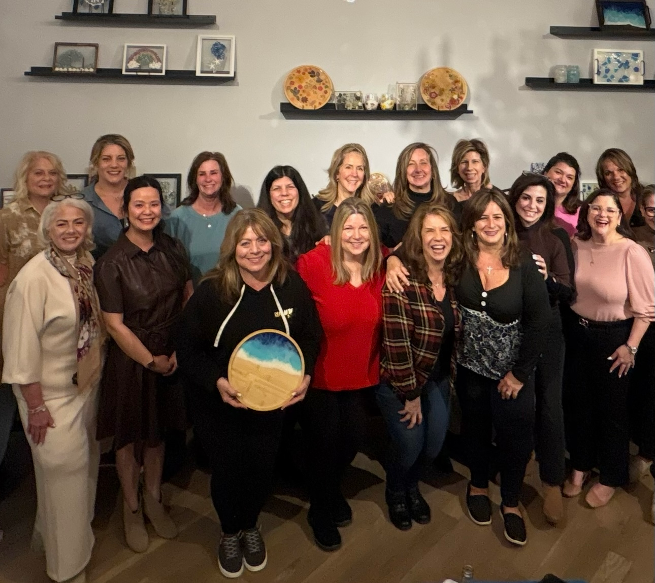Group of smiling people posing indoors, holding a round beach-themed sign with shelves and decor in the background