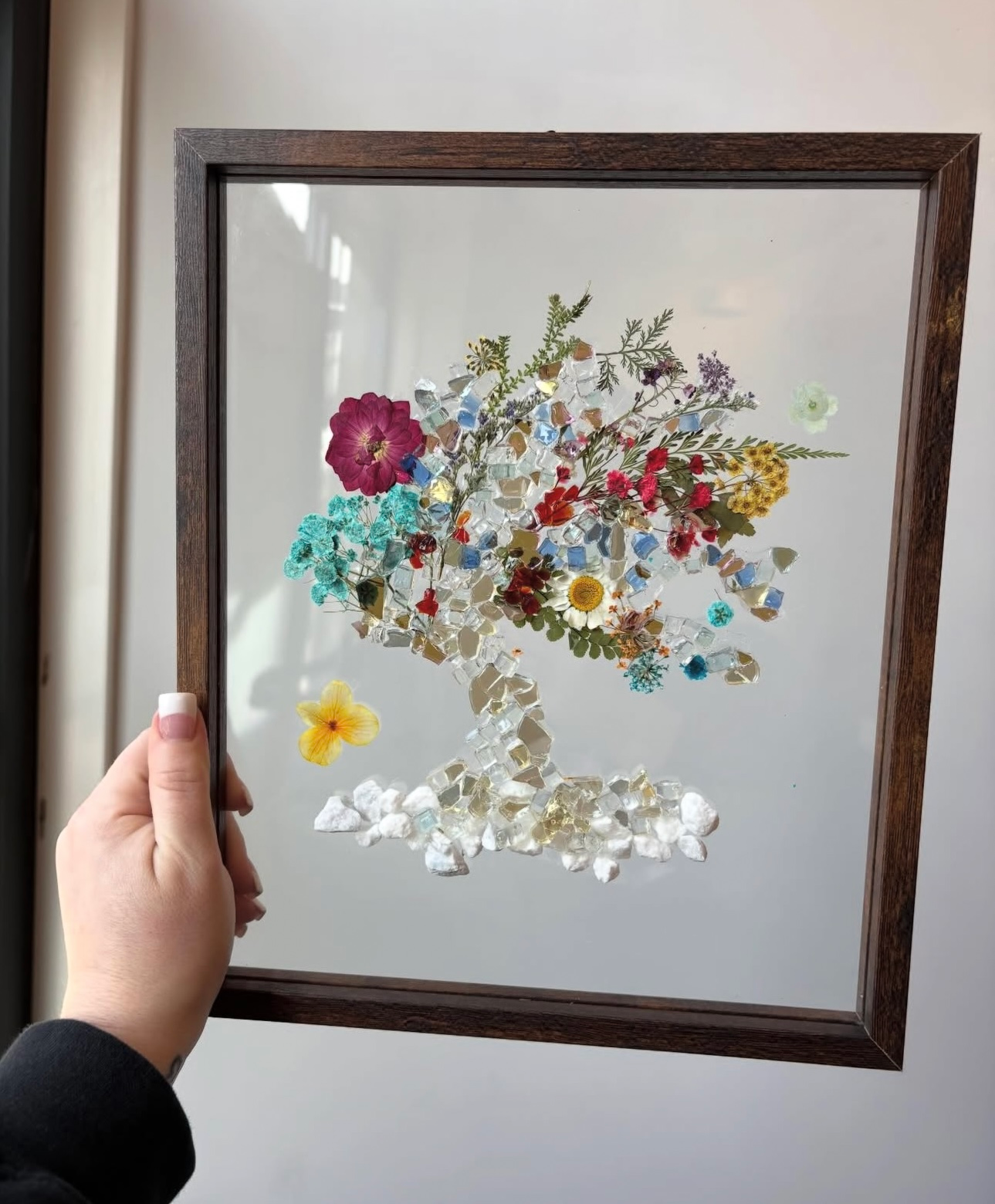 Hand holding framed pressed-flower artwork with colorful floral cluster on a mirrored background