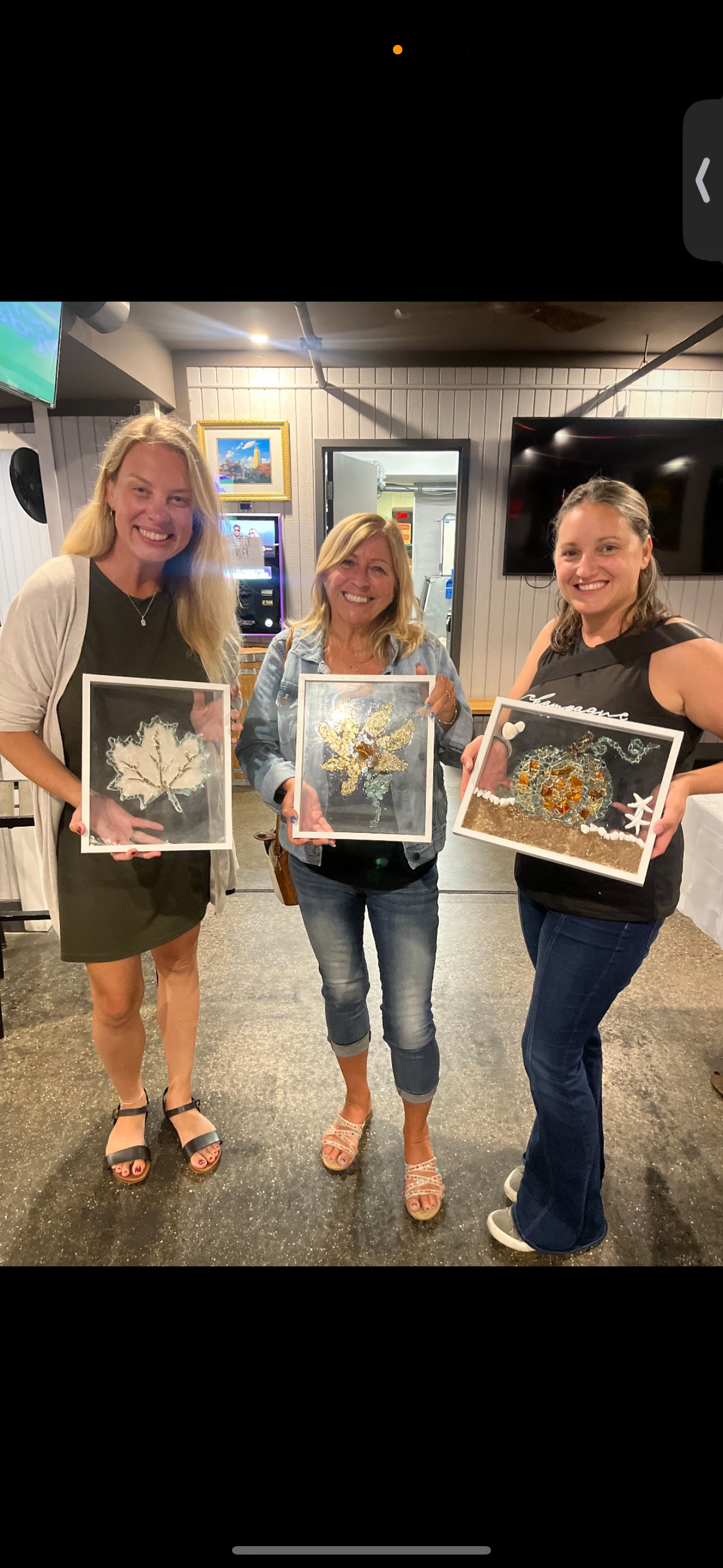 Three women indoors holding framed artwork, smiling in a gallery-like setting.