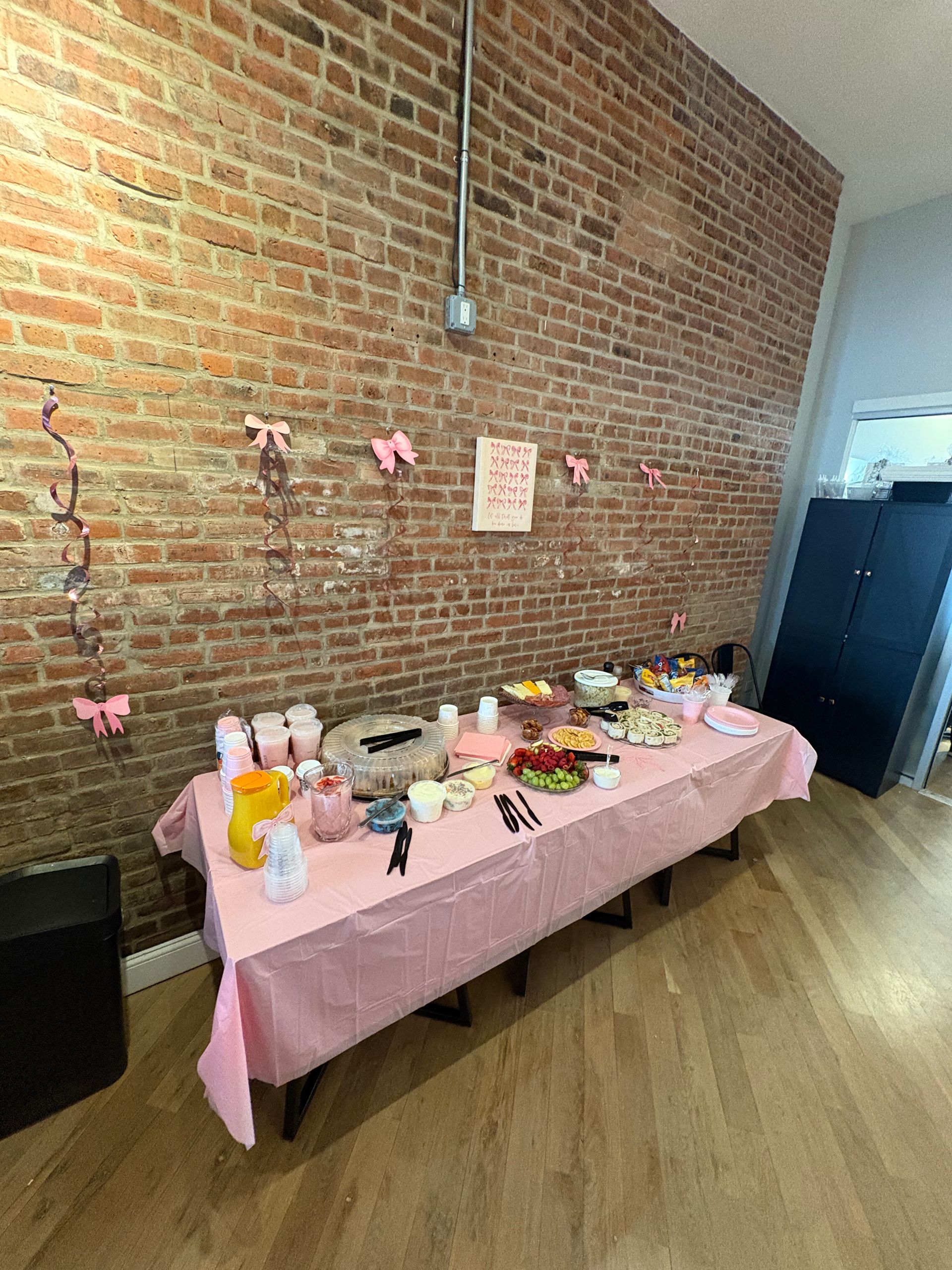 Pink-covered dessert table against brick wall, with treats, decorations, and balloons in a bright room