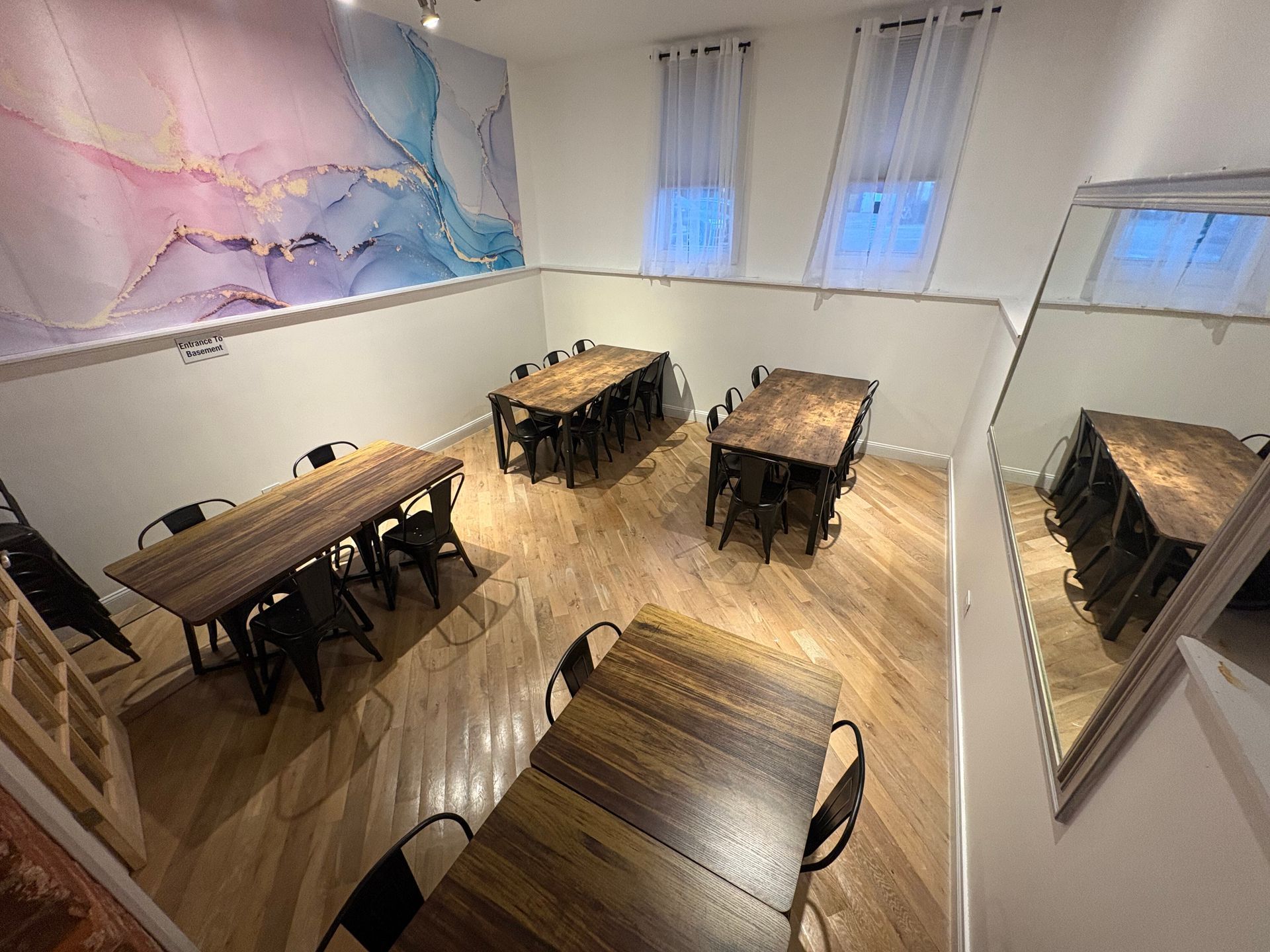 Small café or dining room with wooden tables and black chairs, pastel wall art, and mirrors on the side.