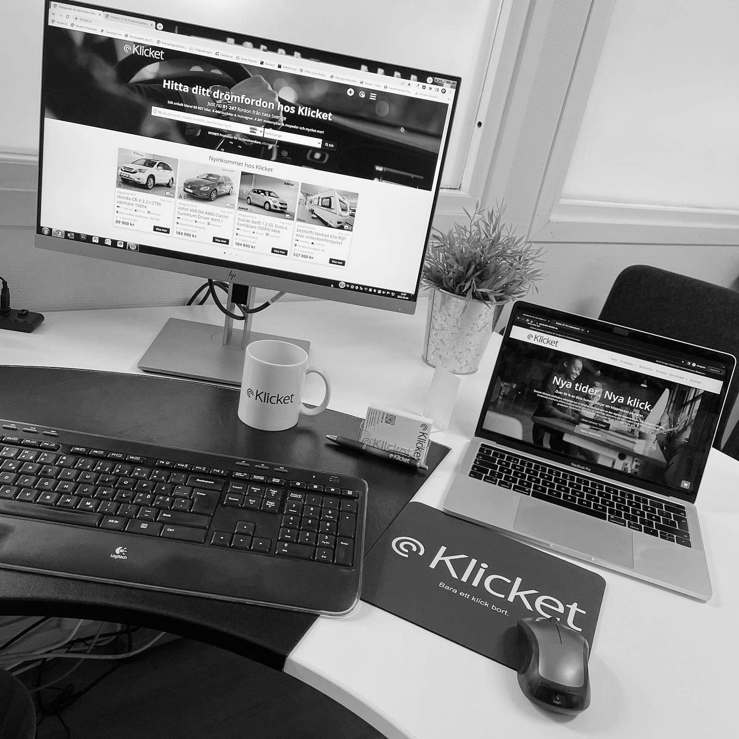 Black and white photo of a desk with a monitor and laptop displaying a website. A keyboard, mug, and mouse are also on the
