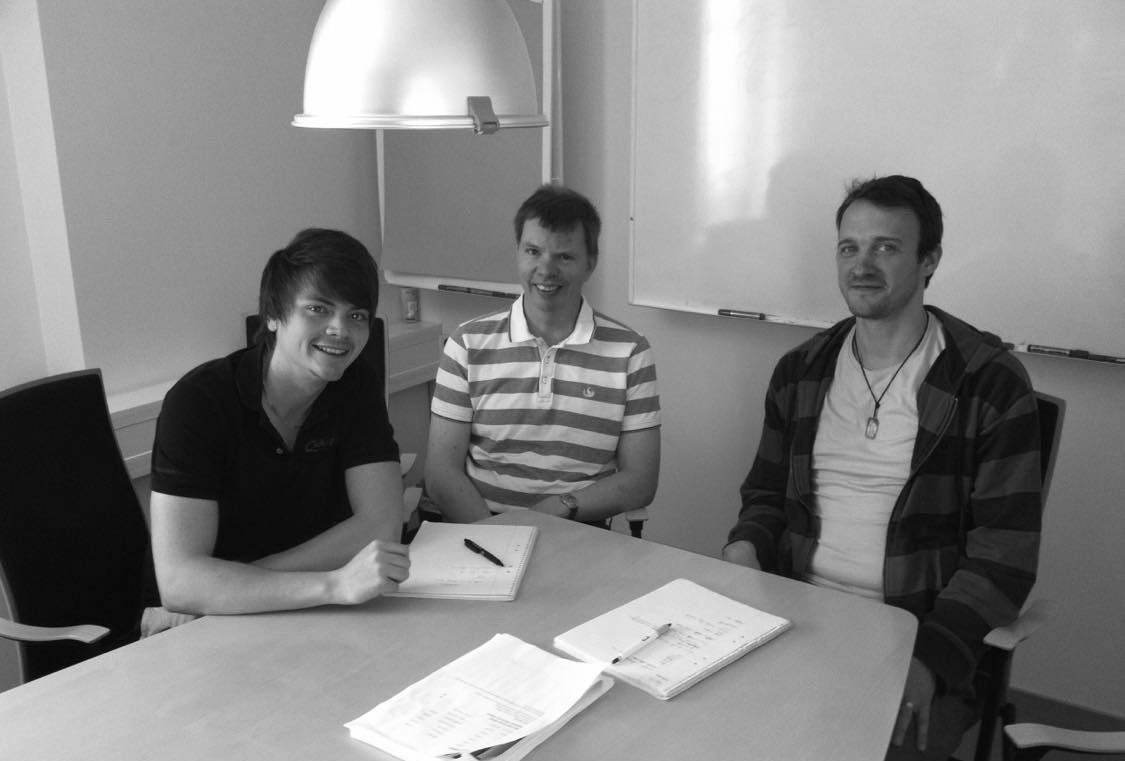 Three people seated around a table with papers, smiling. Bright room, overhead light, whiteboard.