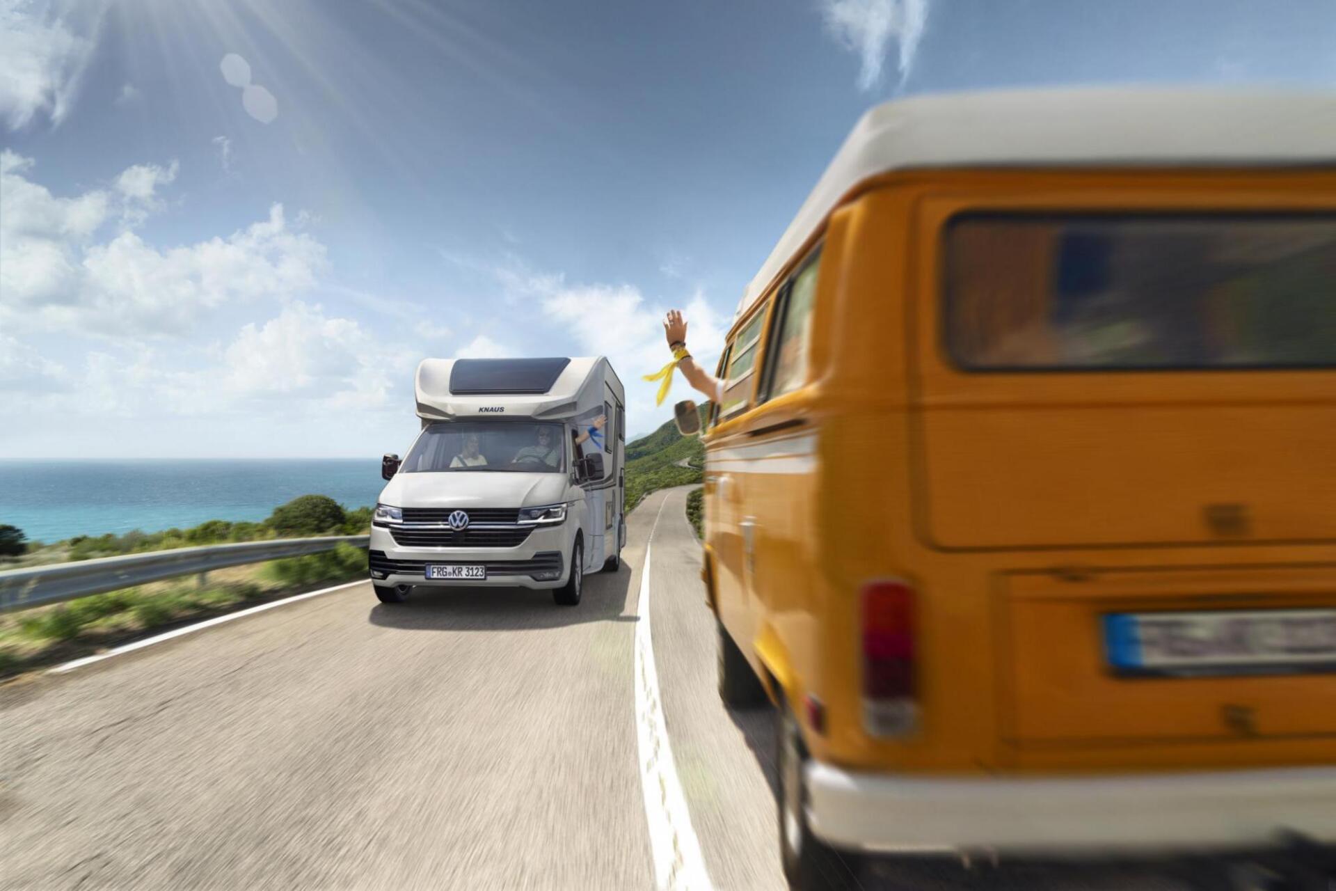 Two camper vans driving on a coastal road; one yellow, the other white. Ocean in the background.