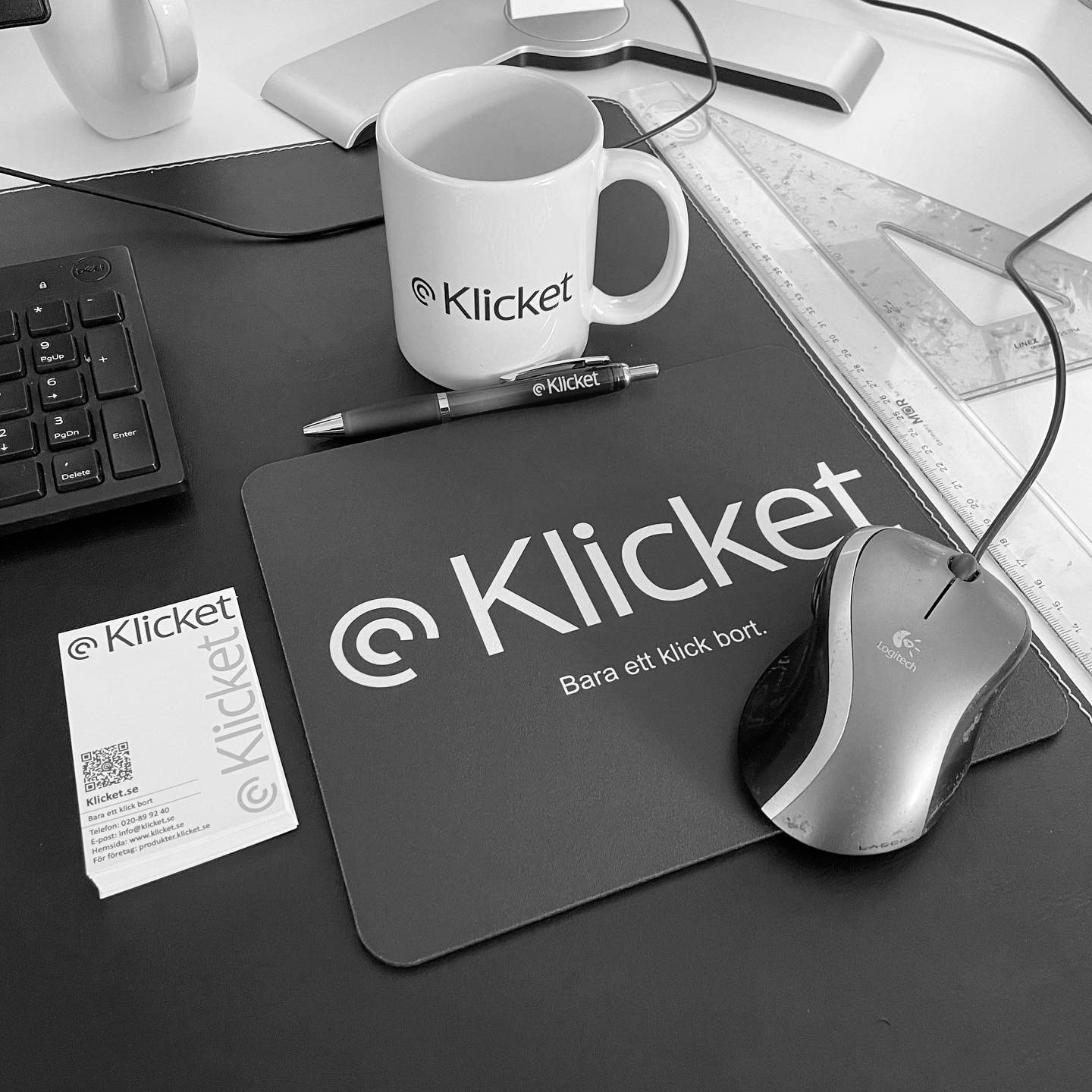 Black and white photo of a desk with a Klicket mug, mousepad, mouse, pen, business card, and calculator.