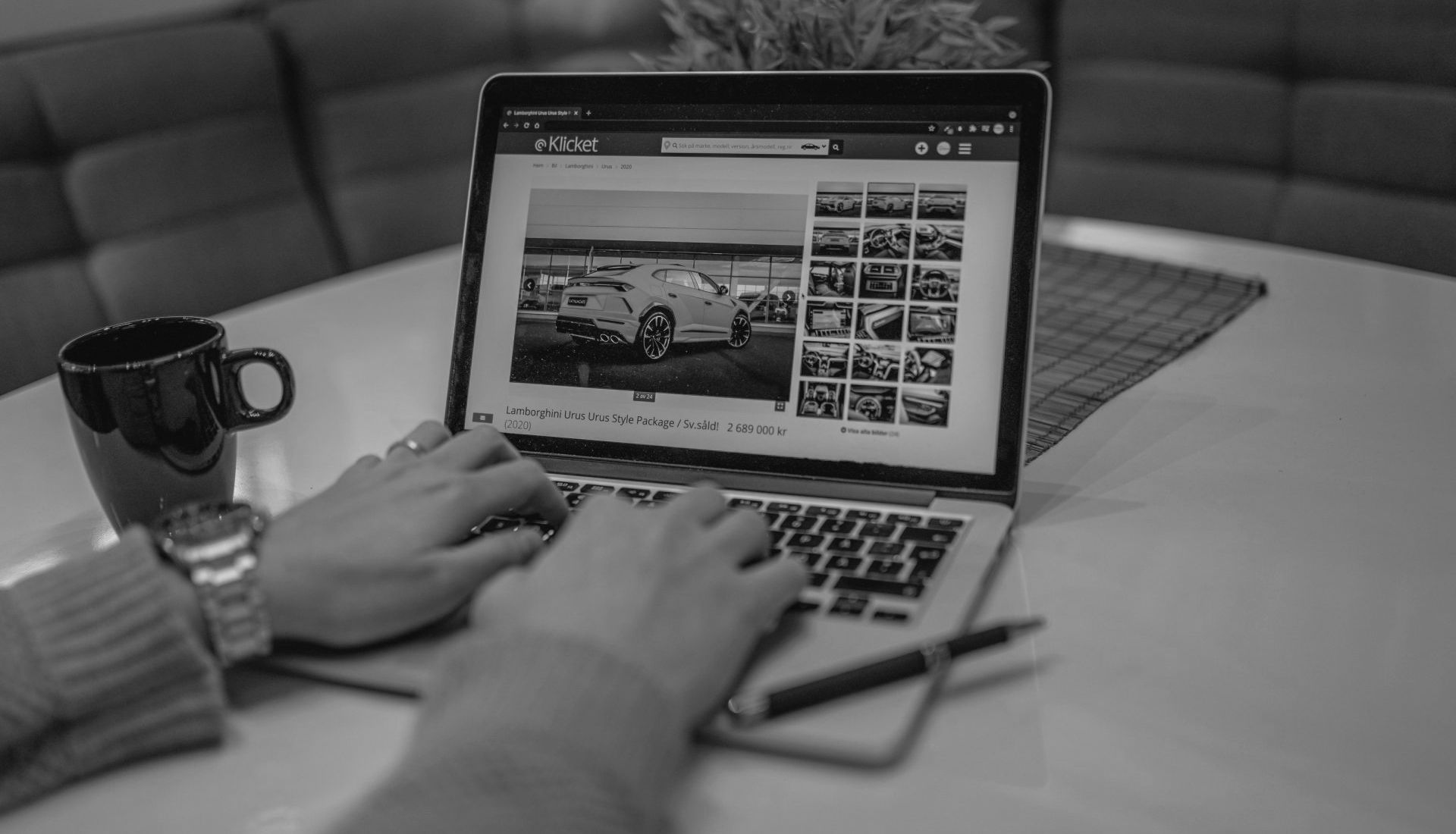 Person typing on laptop, viewing images of a white car. Black mug and pen on table.