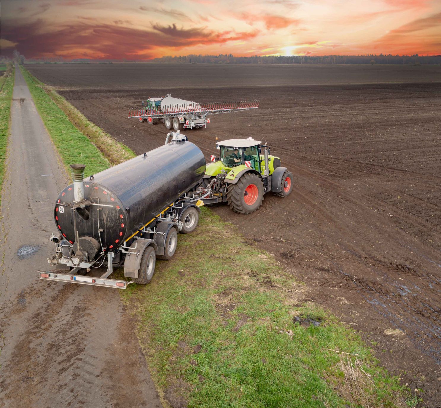 Ein Traktor zieht einen Güllewagen neben einem schlammigen Feldweg bei Sonnenuntergang.