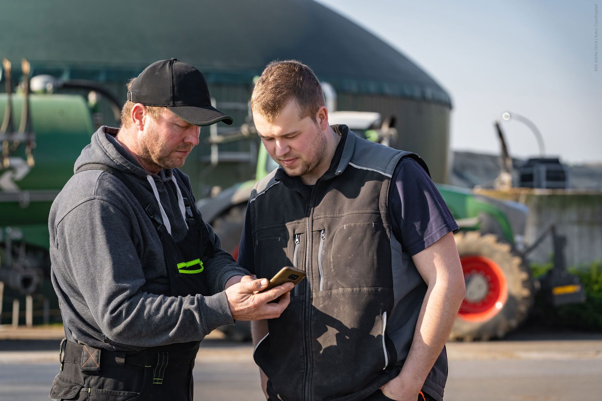 Zwei Männer in Arbeitswesten betrachten im Freien vor einem grünen Traktor ein Smartphone.