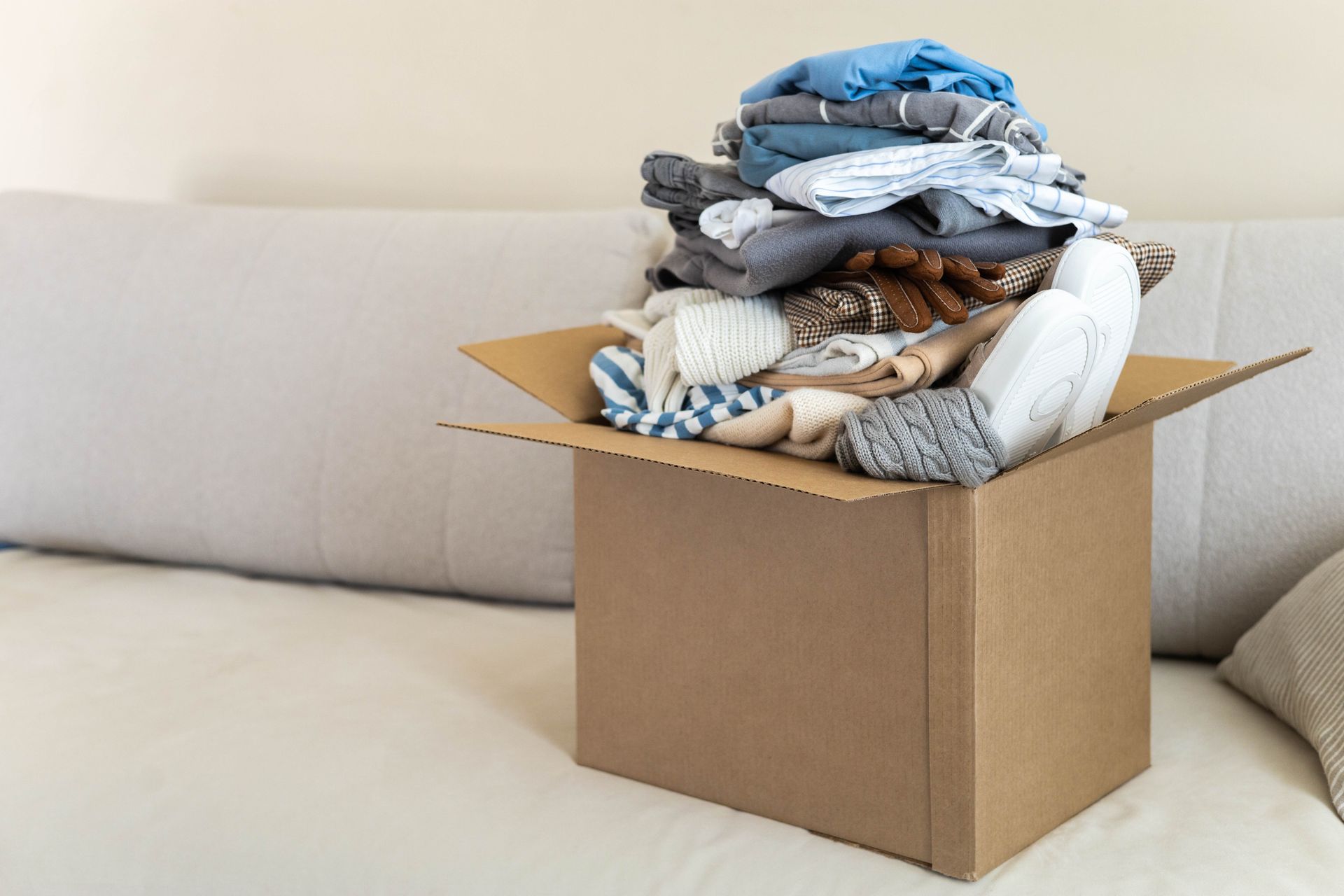 Cardboard box overflowing with folded clothes on a light-colored couch.
