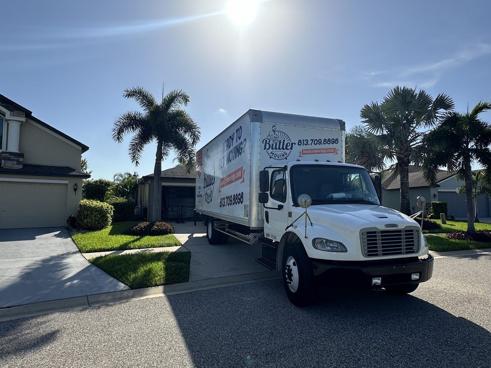 White moving truck parked on a sunny suburban street beside a house and palm trees.