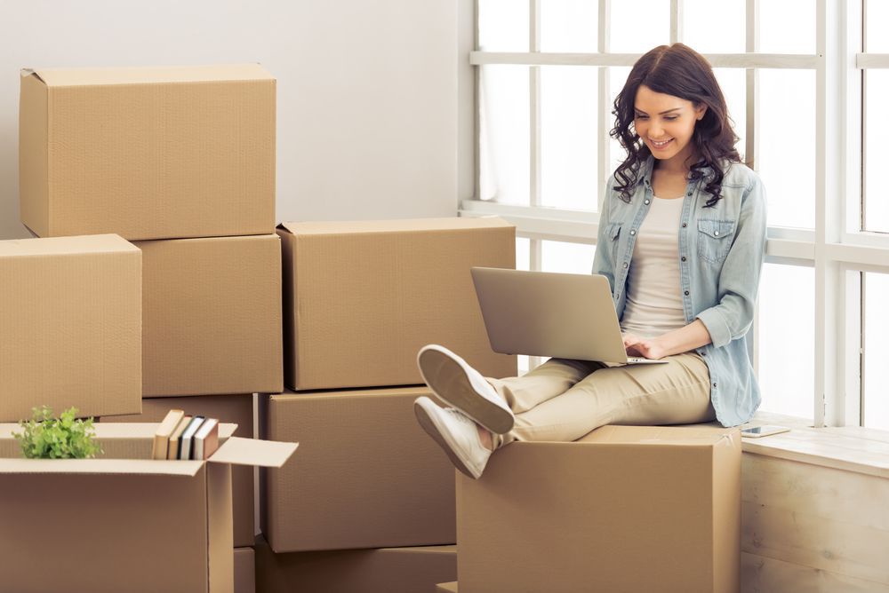 Woman sitting on a moving box with a laptop, surrounded by stacked cardboard boxes in a bright room
