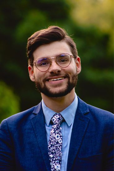 Smiling person in a blue suit jacket and patterned tie, standing outdoors with a blurred green background