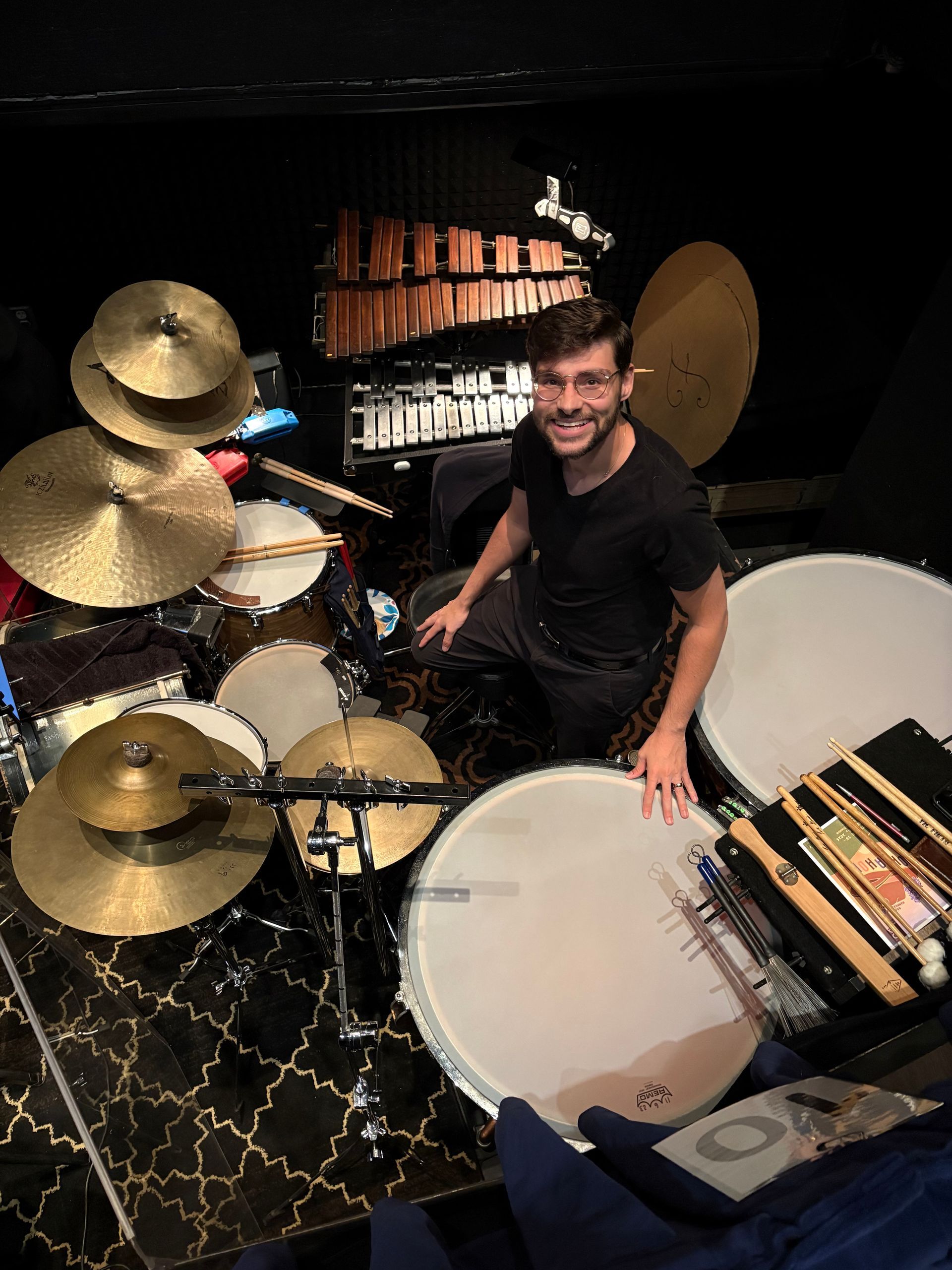 Man smiling beside a drum kit with large cymbals in a dark studio room