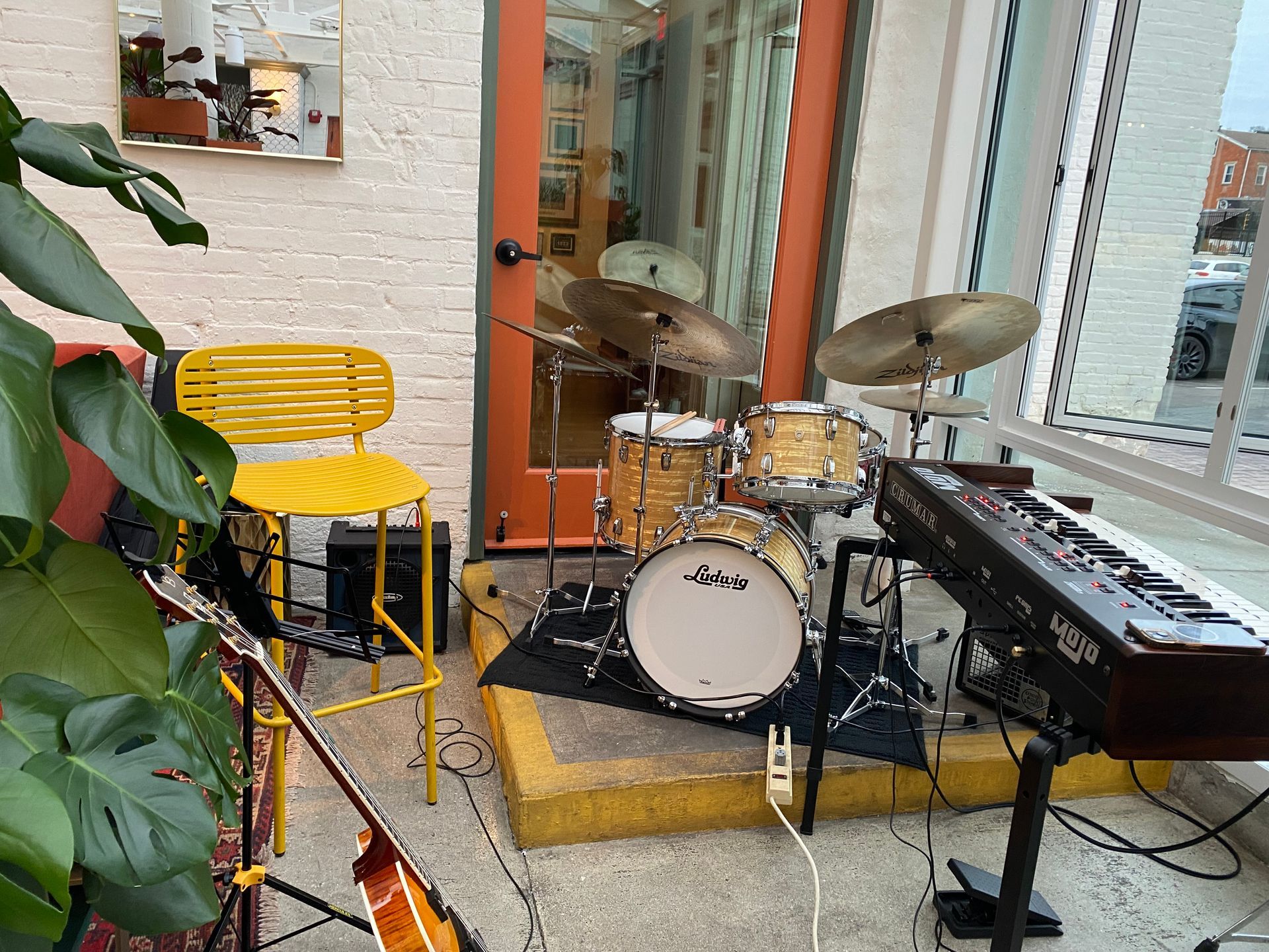 Small music stage with a drum kit, keyboard, yellow chair, and plants beside a window.
