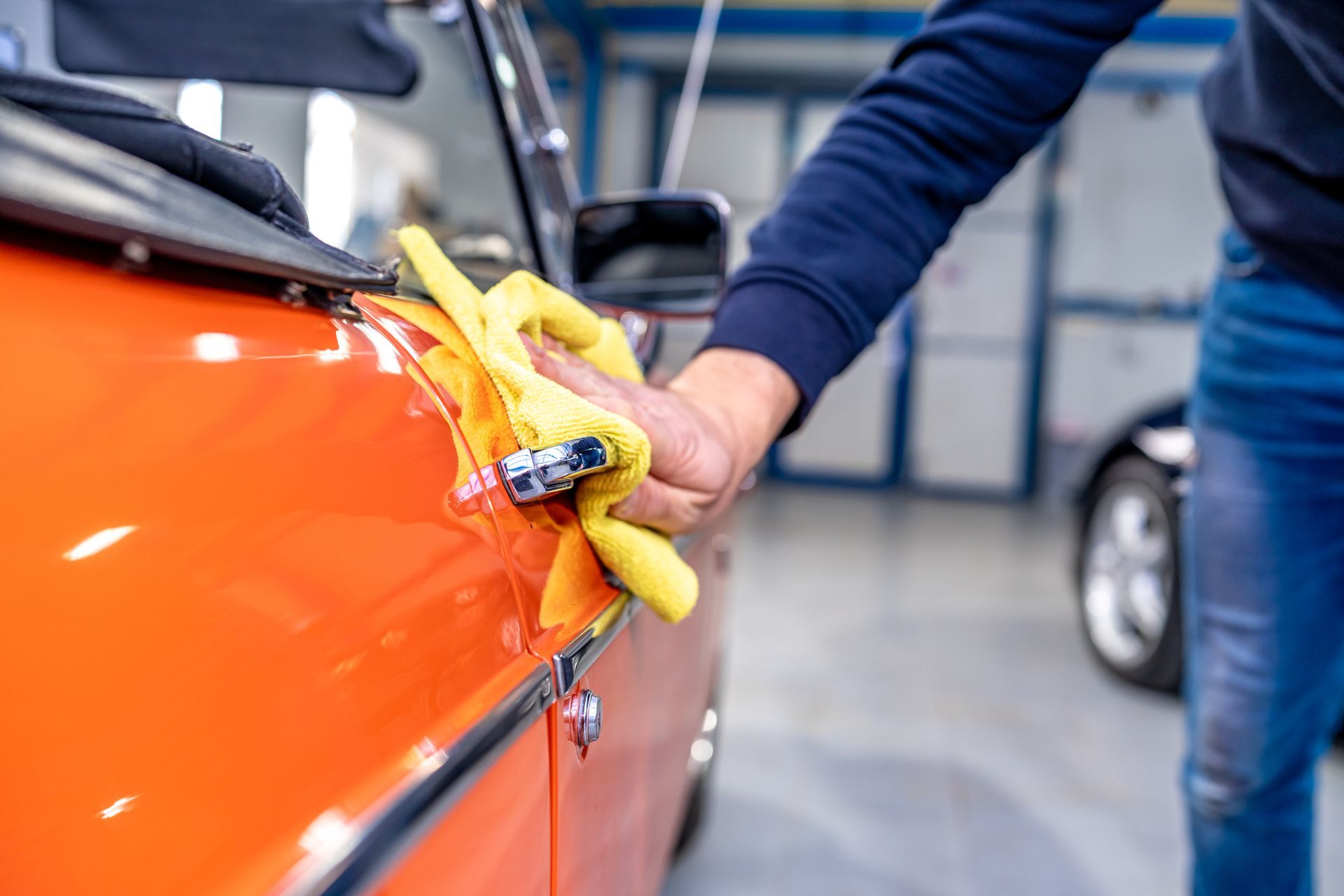 Person wiping down an orange convertible with a yellow cloth in a garage.