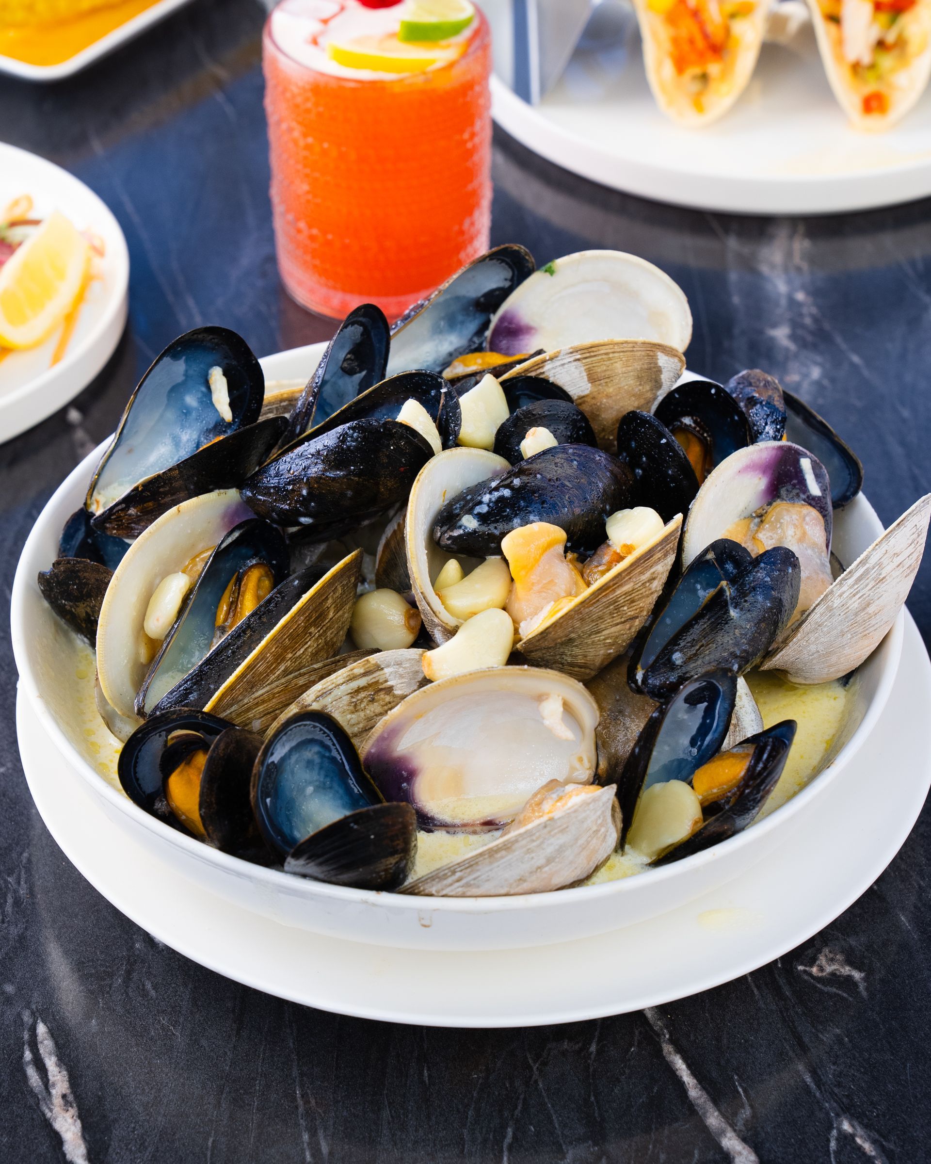 Bowl of mussels and clams in broth, with a cocktail and tacos in the background.