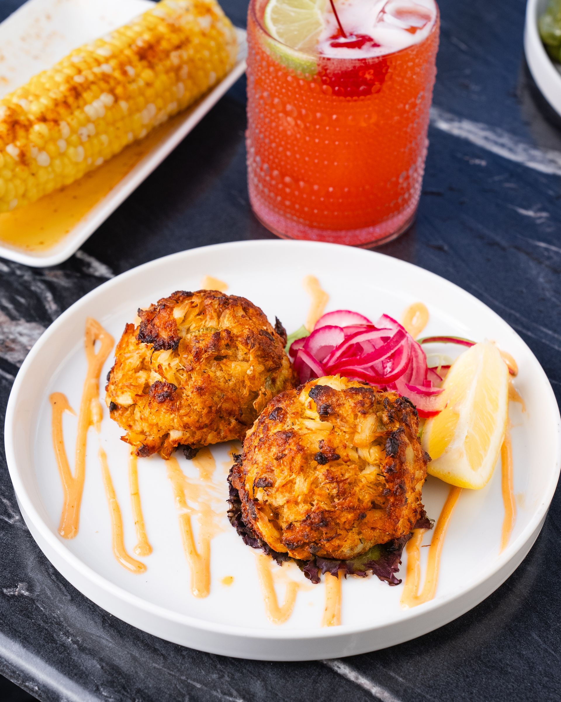 Two crab cakes on a white plate, drizzled with sauce, pickled onions, lemon wedge, and cocktail; corn on the cob