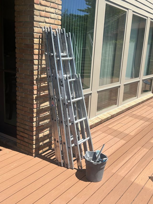Person standing on a ladder indoors, cleaning a tall window in a bright living room.