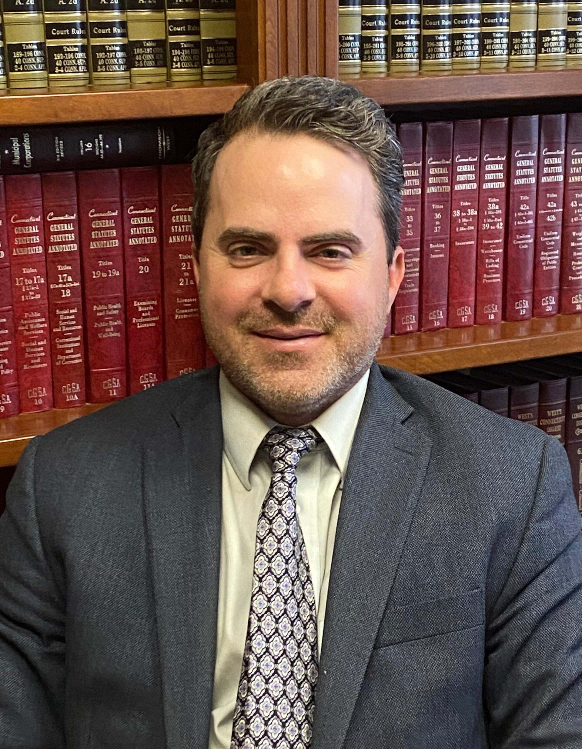 Jonathan Chomick, Attorney — Plainville, CT — Mastrianni & Seguljic LLC Man in suit, tie, smiling, in front of a bookshelf filled with law books.