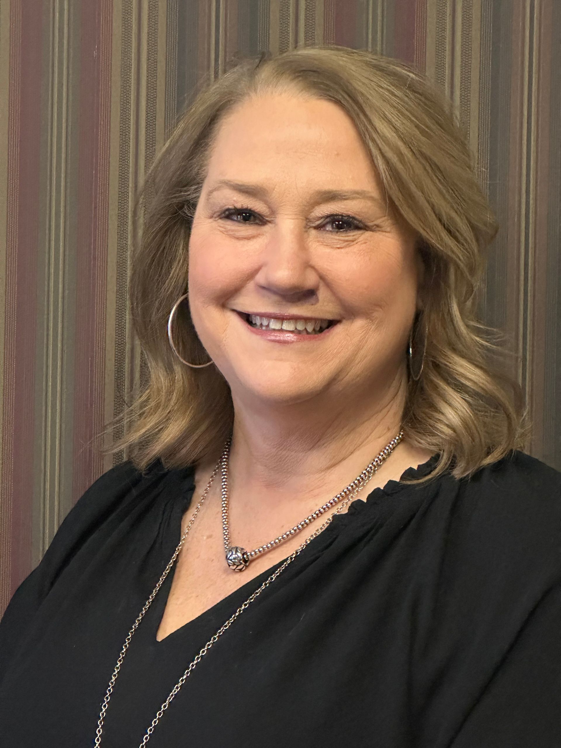 Tami Lynne Pinardi — Plainville, CT — Mastrianni & Seguljic LLC Woman with blonde hair smiling, wearing black top and silver necklace, against striped background.
