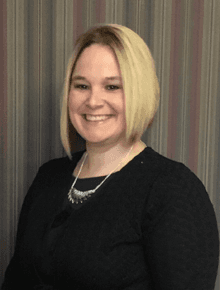 Stephanie Gibbons, Paralegal — Plainville, CT — Mastrianni & Seguljic LLC Woman with blonde bob smiles, wearing a black sweater and silver necklace, in front of striped background.