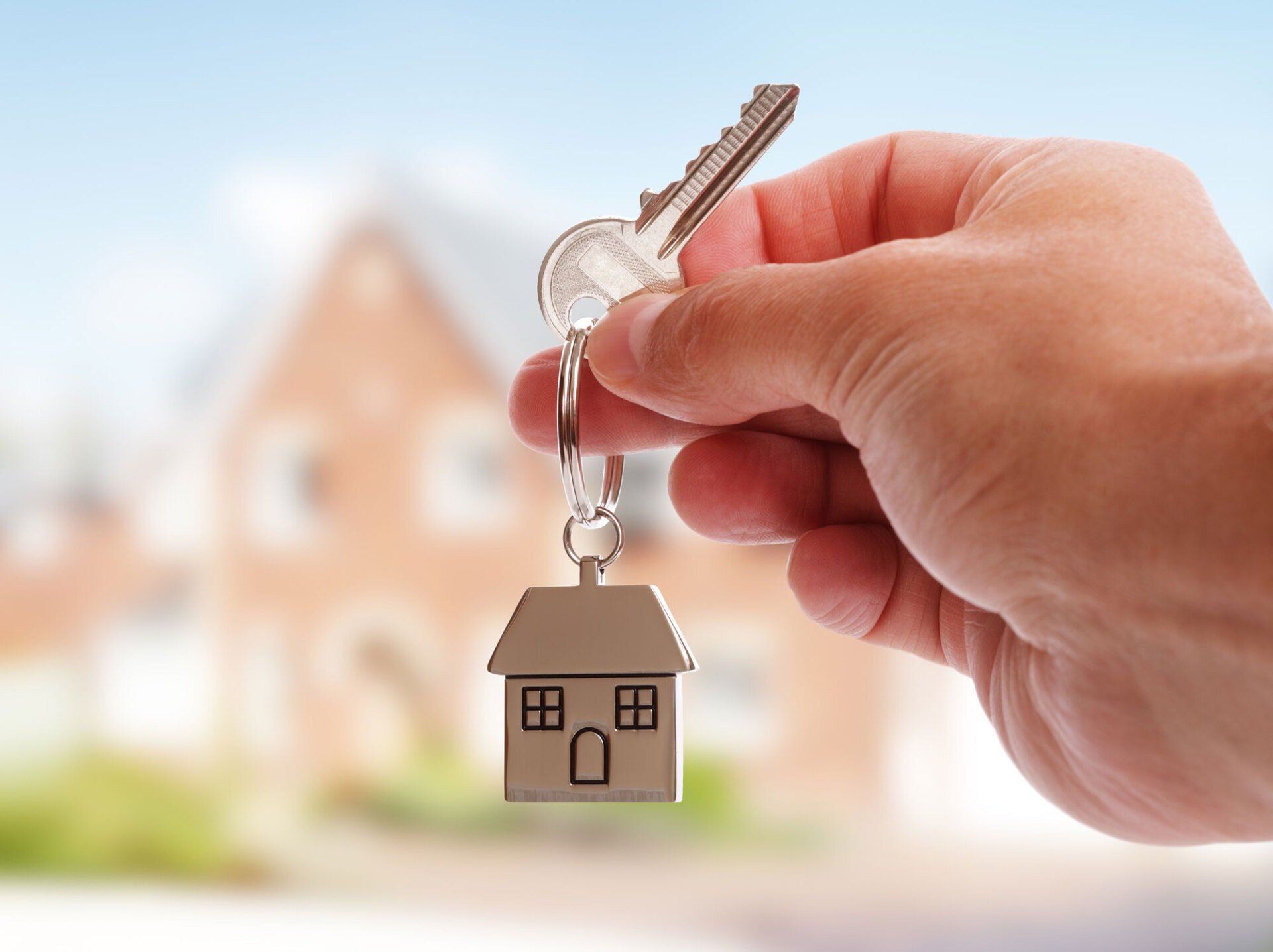 Hand holding house key with a keychain house, in front of a blurred brick house.