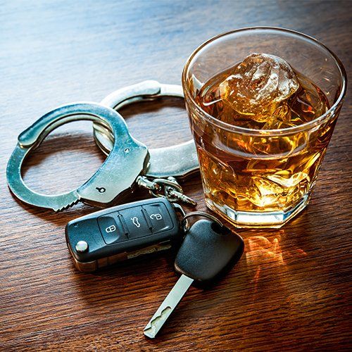 Whiskey with Car Keys and Handcuffs — Plainville, CT — Mastrianni & Seguljic LLC Handcuffs, car keys, and a glass of whiskey with ice on a wooden table.