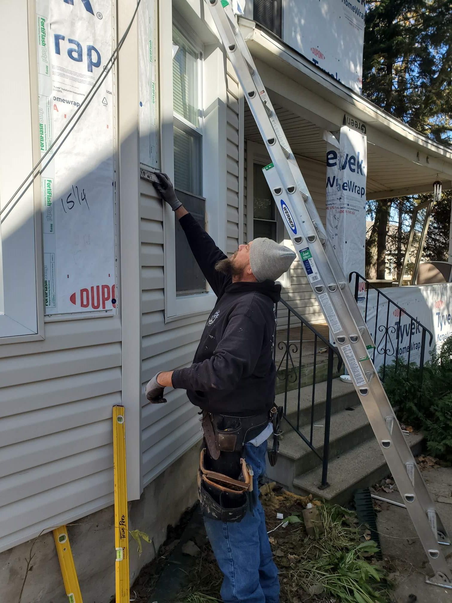 Worker on a ladder inspecting house siding beside a window and porch.