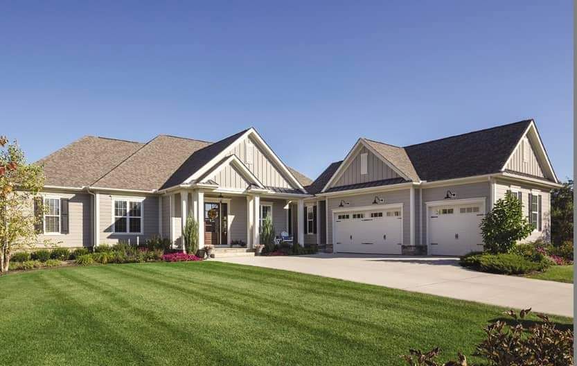 Single-story suburban house with beige siding, gabled roofs, two-car garage, and a green lawn under a blue sky