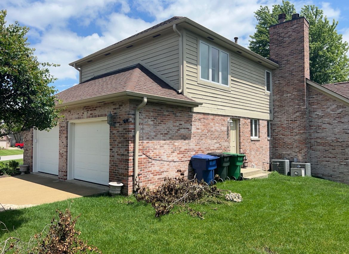 Suburban brick-and-siding house exterior with two garages, chimney, lawn, and blue recycling bins
