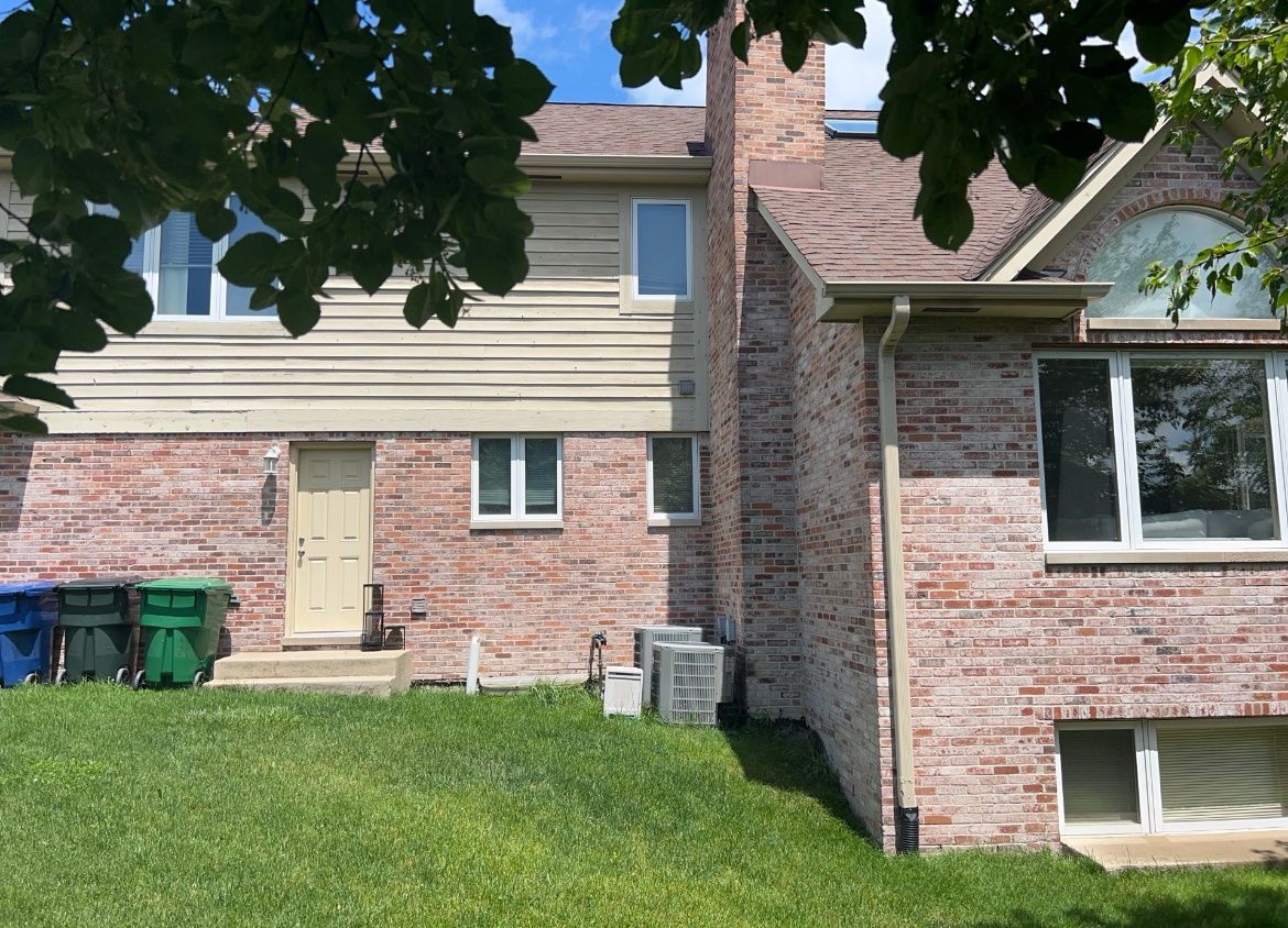 Brick house exterior with tan siding, green trash bins, and a grassy front yard under tree shade
