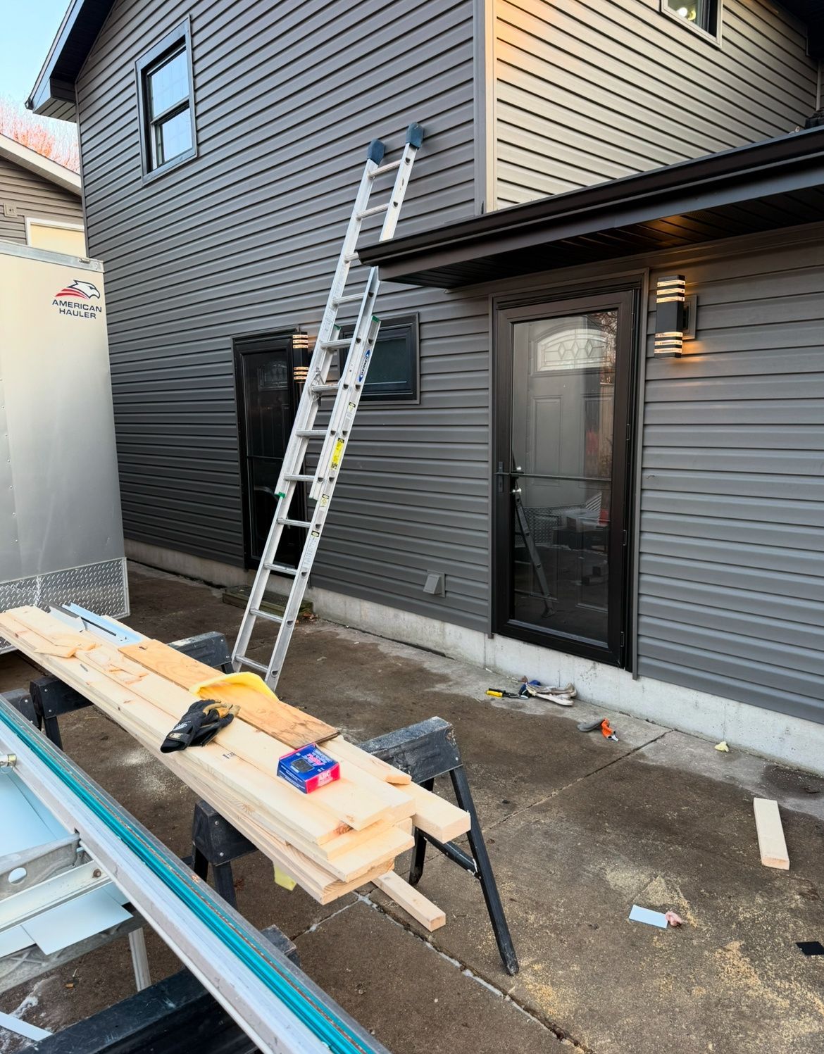 Ladder leaning against gray house exterior beside glass door; lumber and tools on sawhorses in foreground.